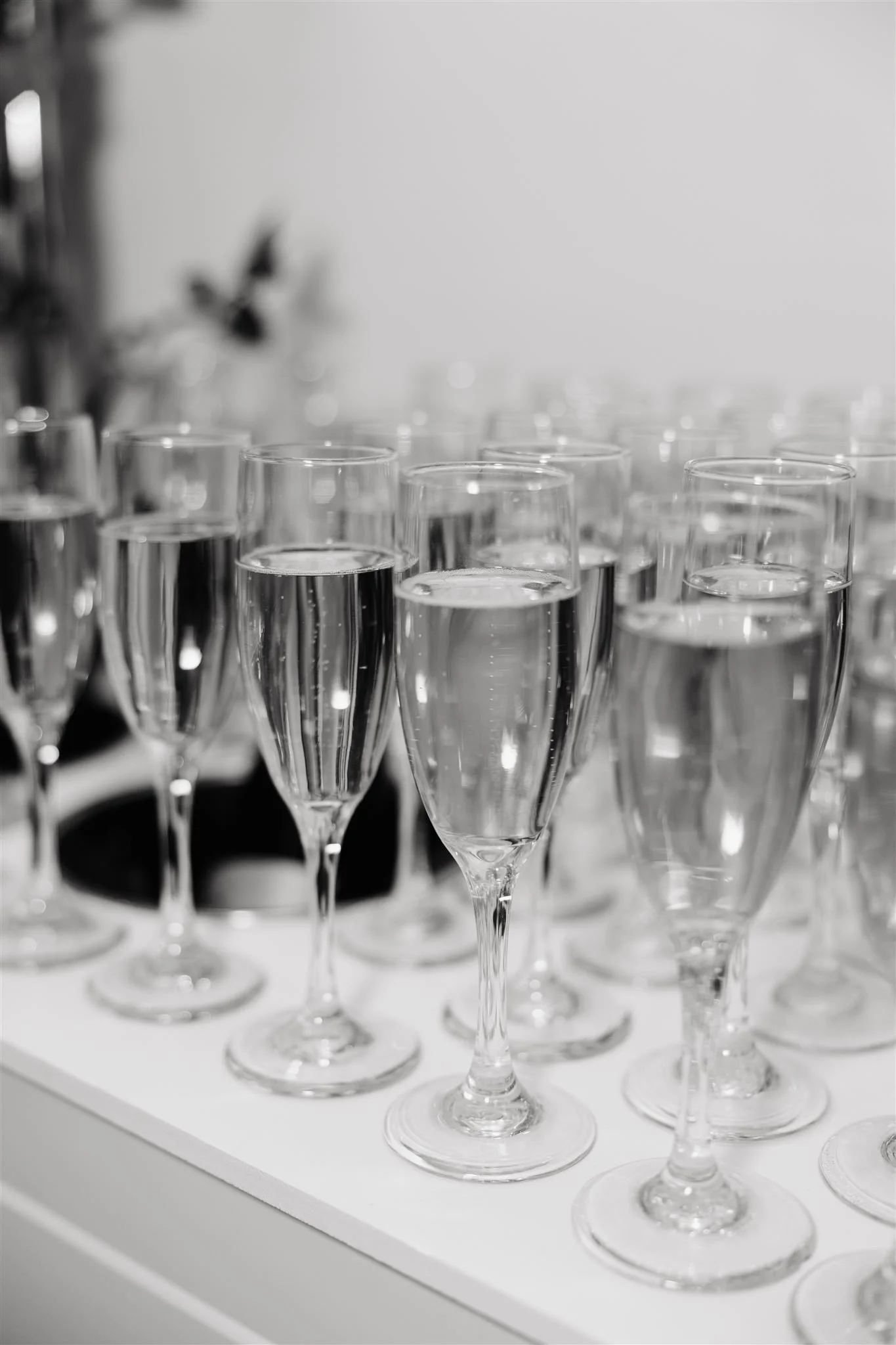 Multiple champagne glasses filled with clear liquid arranged on a white surface, with a blurred background.