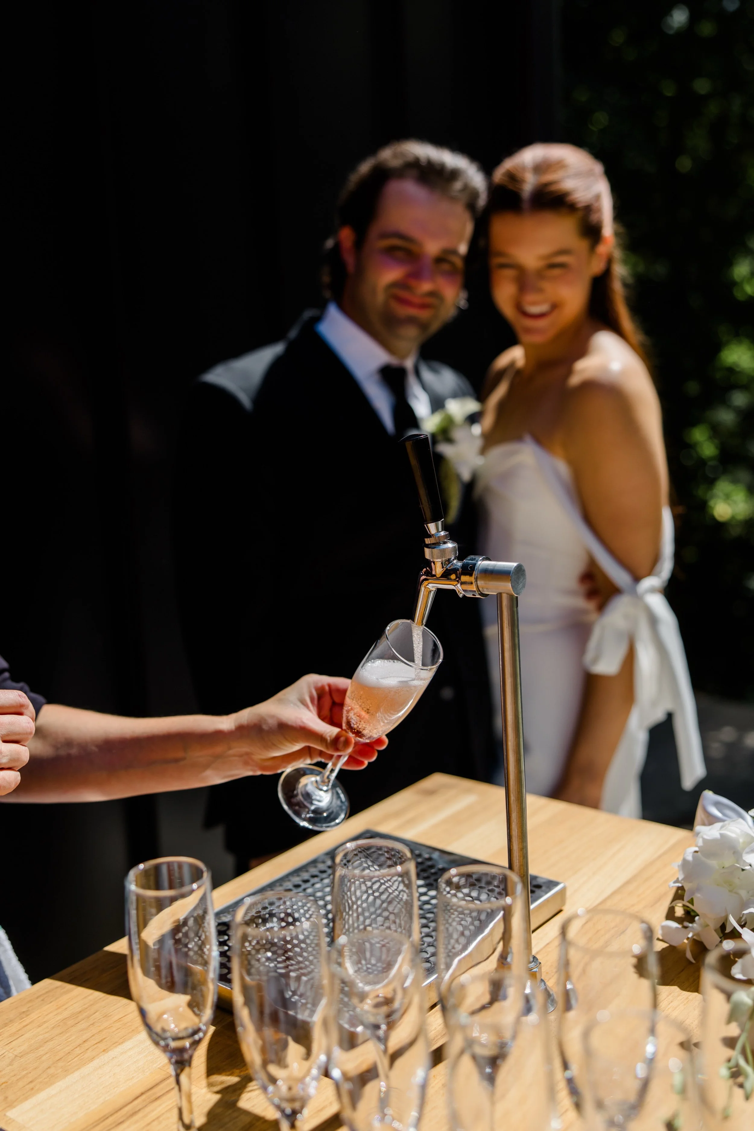 A bride and groom smiling in the background, with a person pouring champagne into a glass from a tap at a wedding celebration.