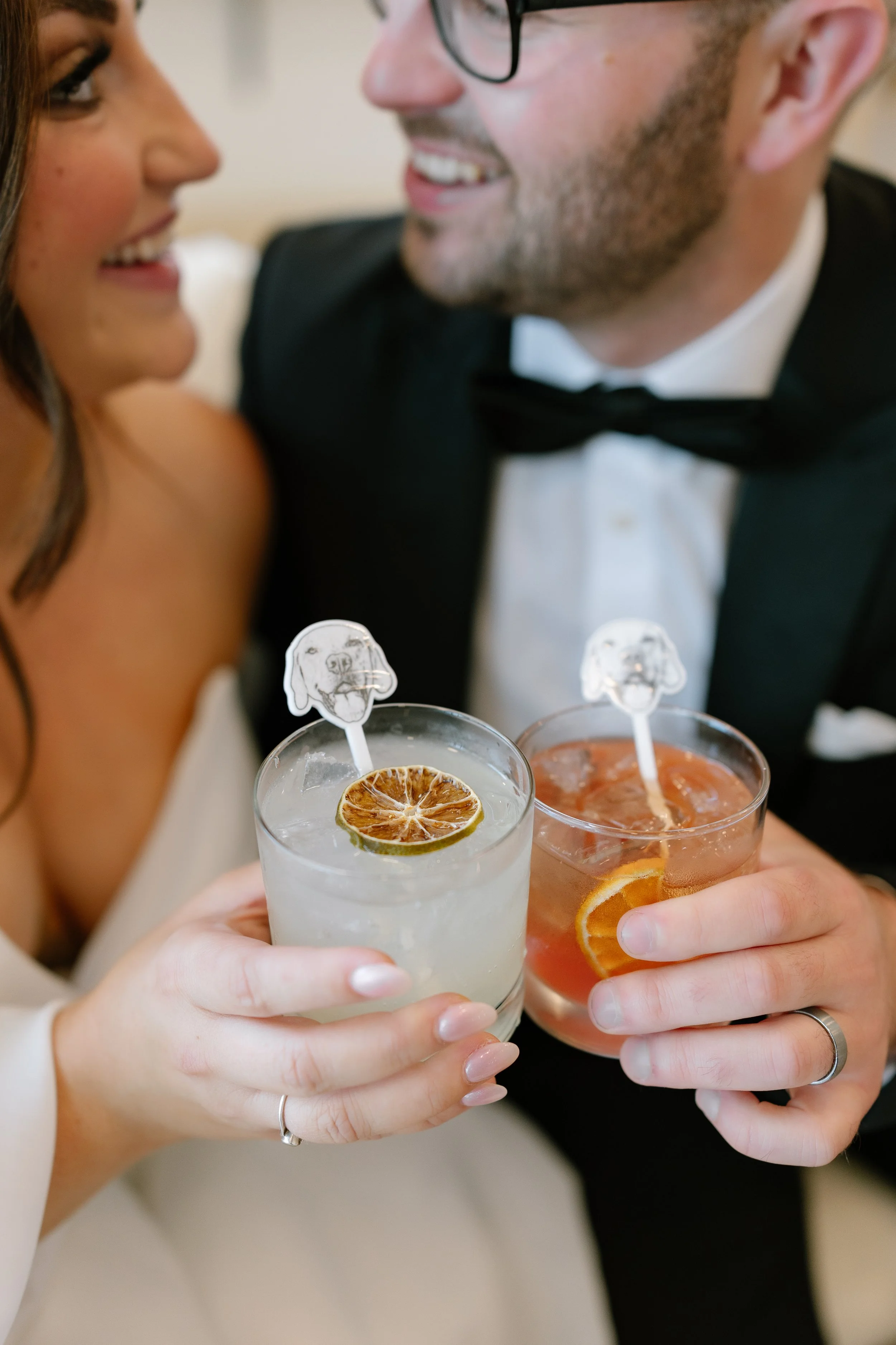 A bride and groom in wedding attire holding custom cocktails with custom drink stirrers, smiling and looking at each other.