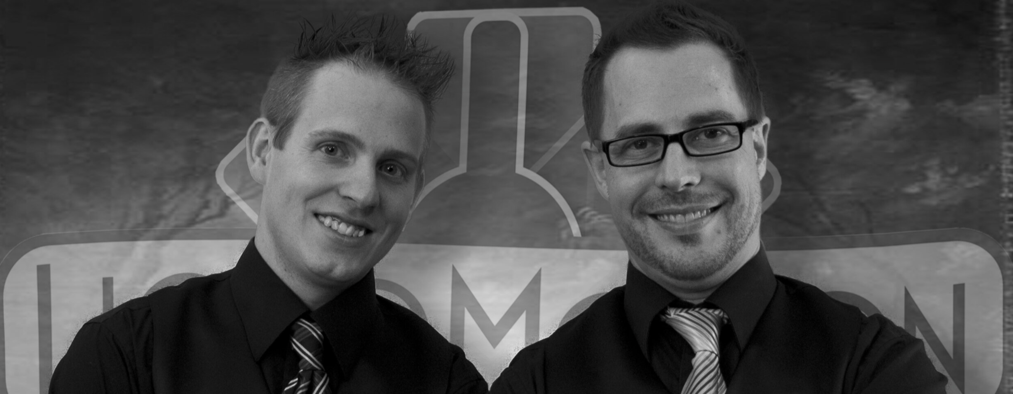 Two men in dress shirts and ties smiling for a photo in front of a sign with a dark background.
