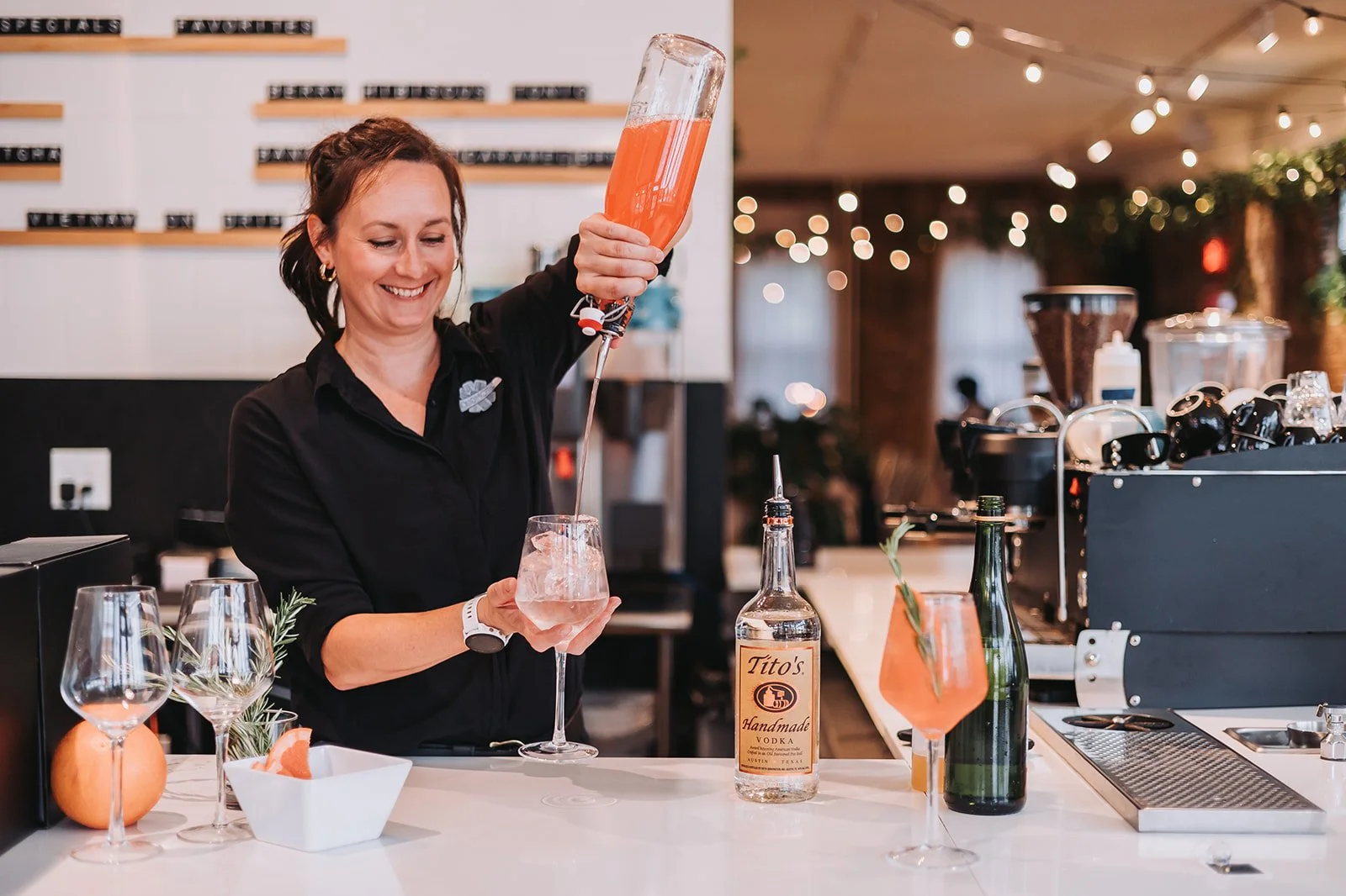 A woman smiling and pouring a pink cocktail into a glass at a bar counter with bottles, glasses, and bar tools, in a warmly lit restaurant or bar setting.