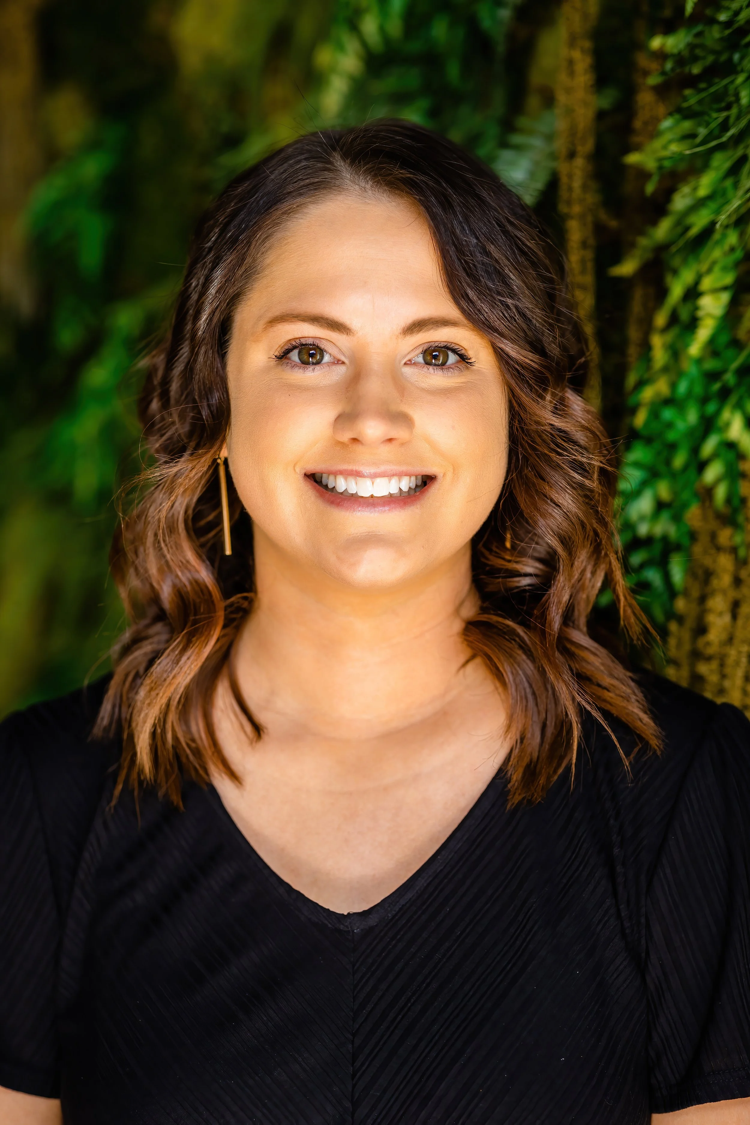 A woman with wavy brown hair smiling in front of a leafy green background.