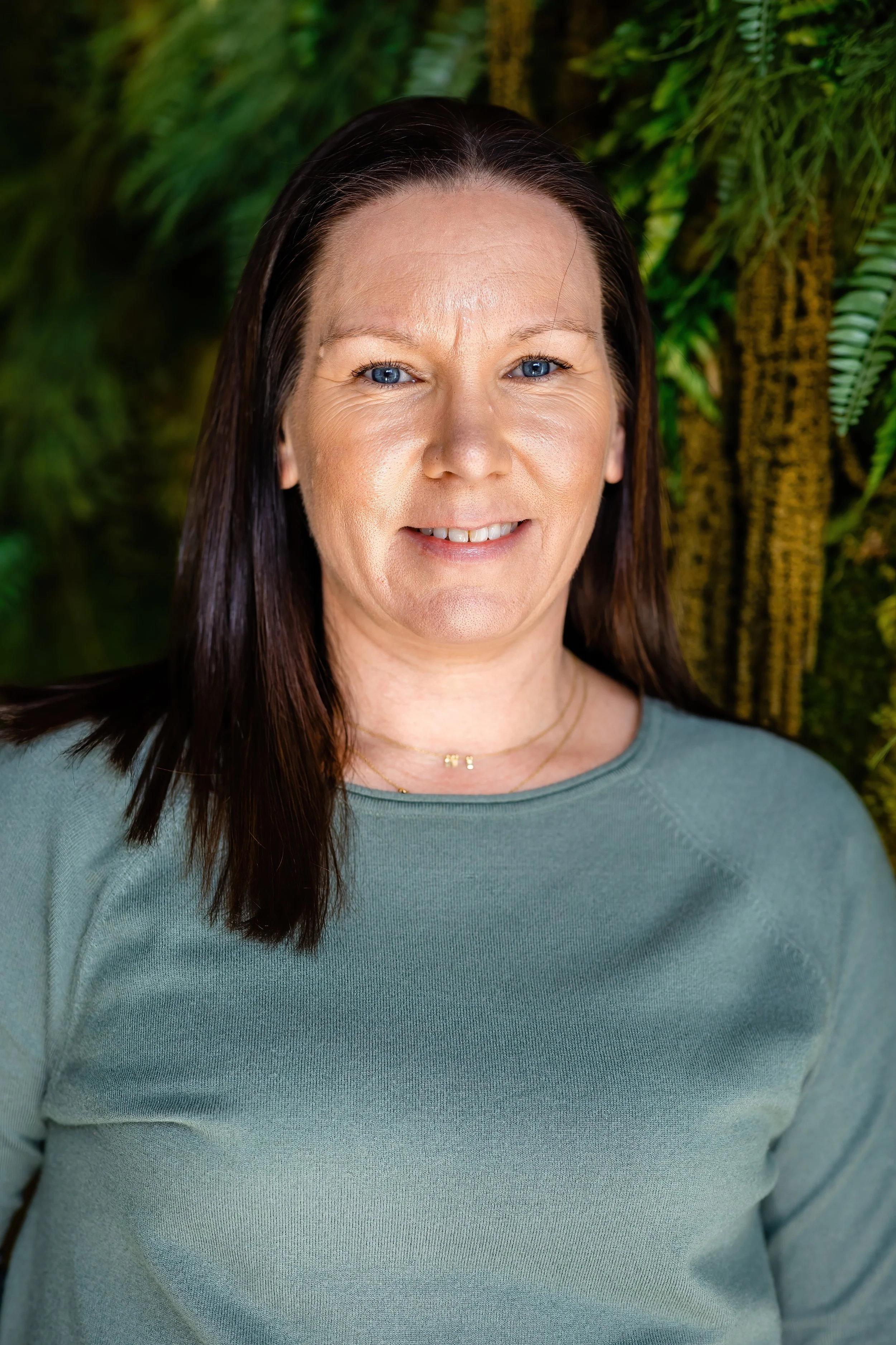 A woman with brown hair and blue eyes smiling at the camera, wearing a green top, set against a background of green ferns.