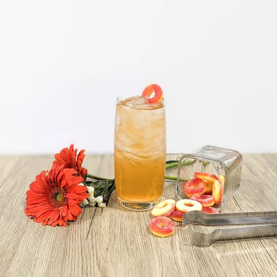 A tall glass of iced tea with a red and yellow gummy ring on the rim, placed on a wooden surface. Beside the glass, there is a overturned container spilling out red and yellow gummy rings, with some on the surface. An orange gerbera daisy flower and small white flowers are also on the surface.