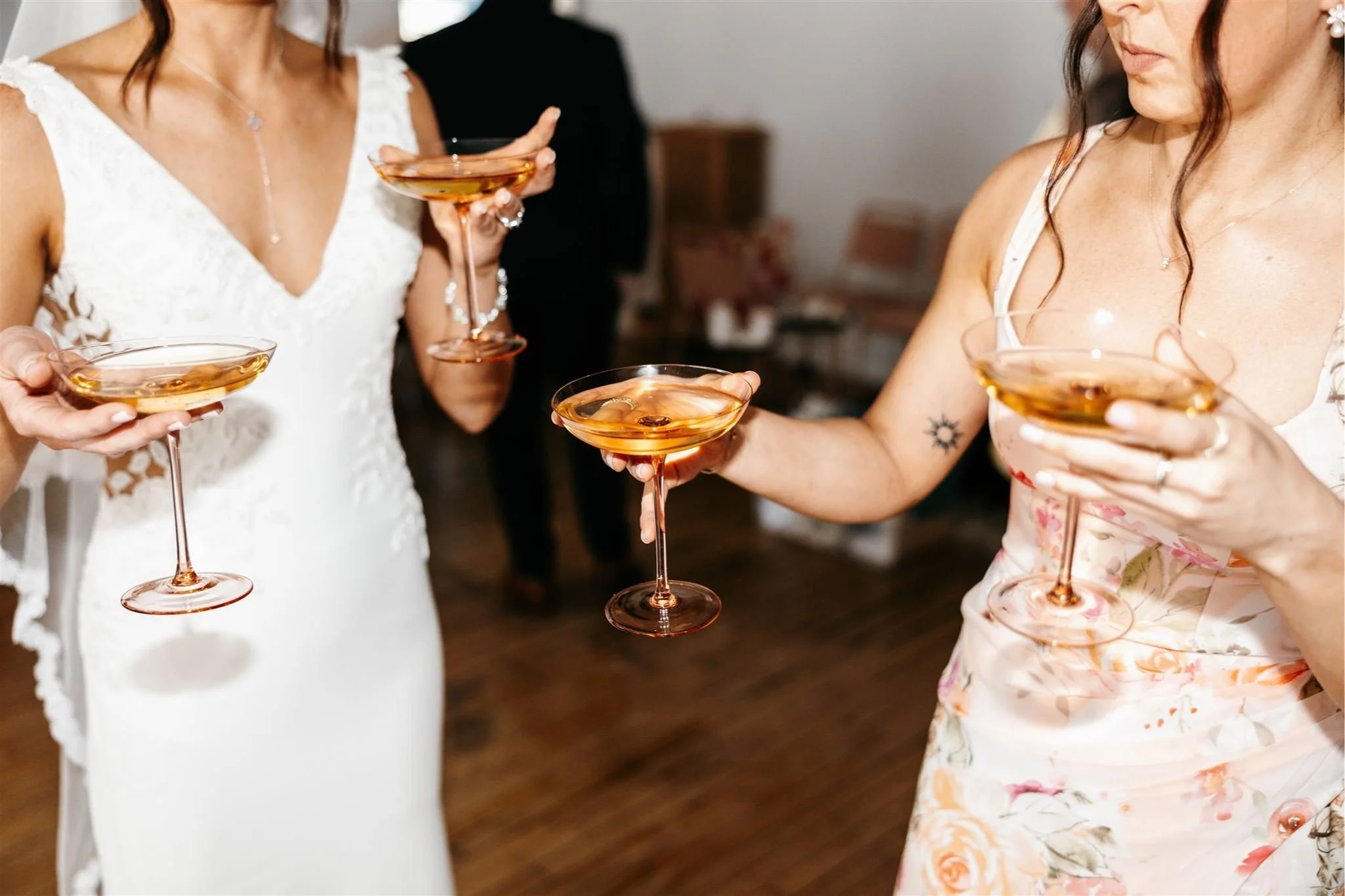 Two women holding champagne coupe glasses with rose-colored drink, standing indoors, one in a white lace dress and the other in a floral dress.