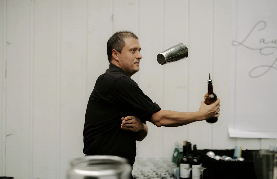 A man in a black shirt is swinging a bottle while a metal cup is falling in front of him.