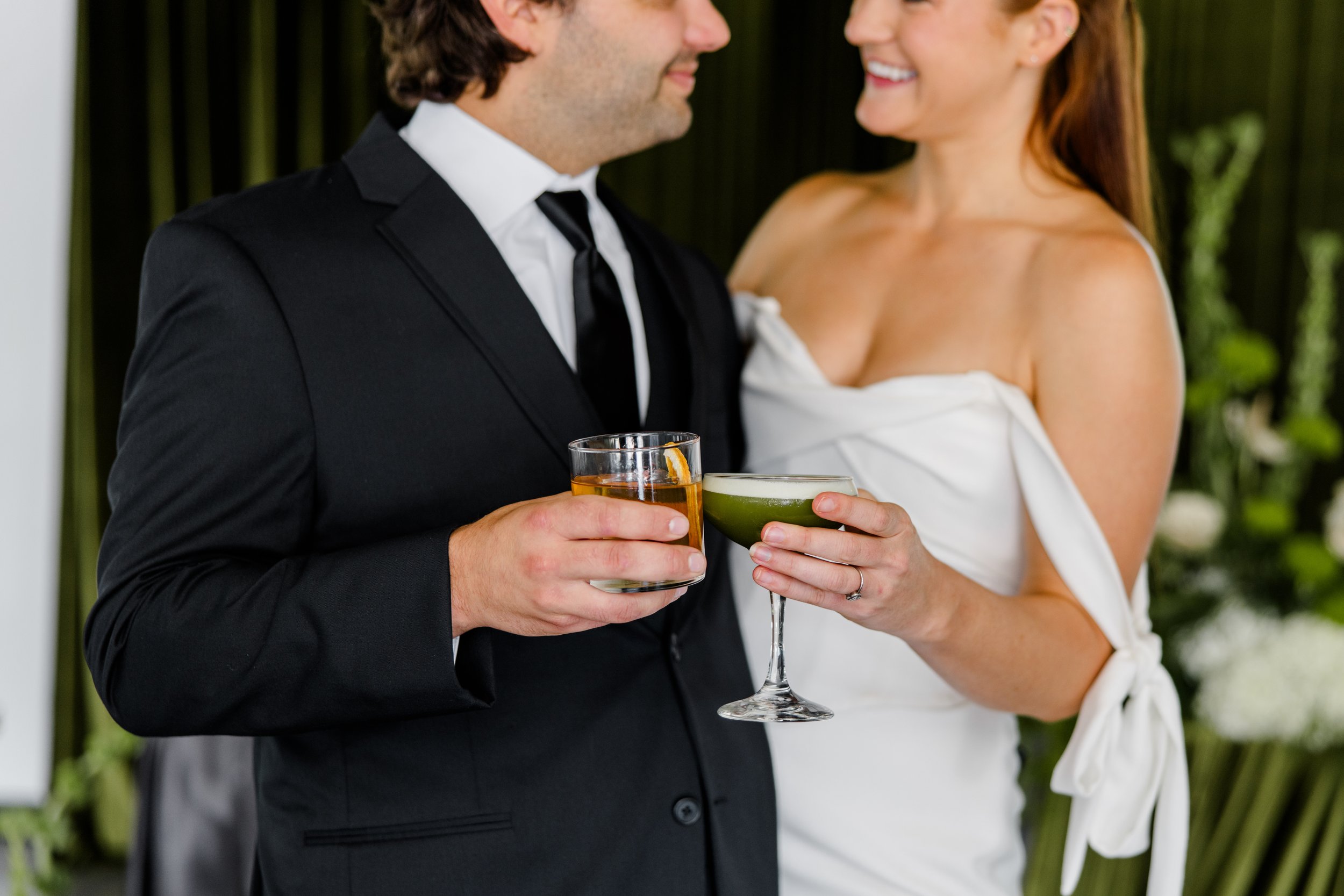 A man and woman dressed formally holding drinks at a celebration, smiling and looking at each other, with blurred greenery in the background.