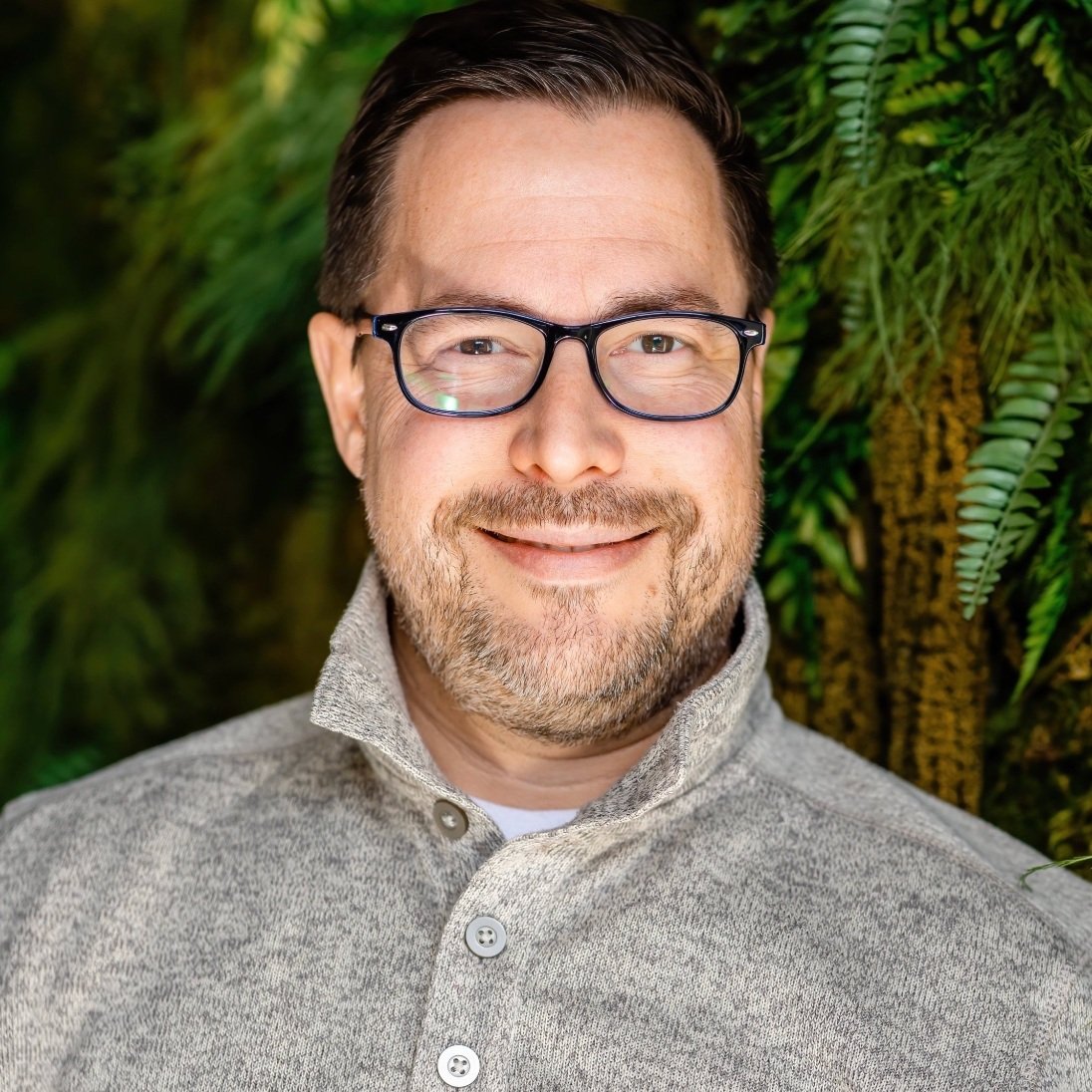 A man with glasses, smiling, wearing a light gray collared sweater, standing in front of green foliage.