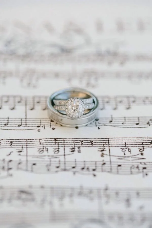 A pair of wedding rings, including an engagement ring with a large round diamond surrounded by smaller diamonds, resting on a sheet of musical notation.