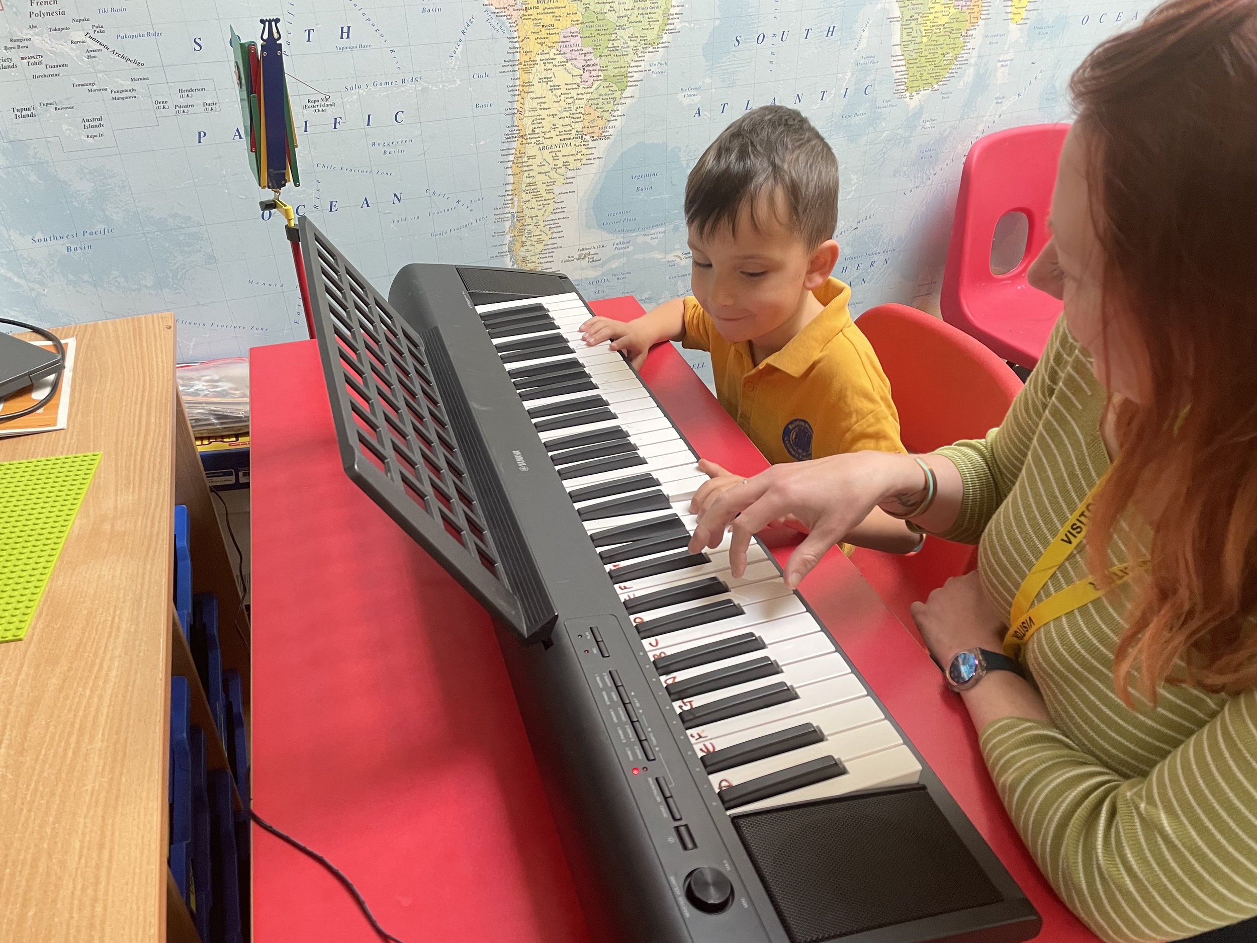 Two young girls with curly red hair playing piano together, sitting on a bench at a black grand piano in a well-lit room near a window.