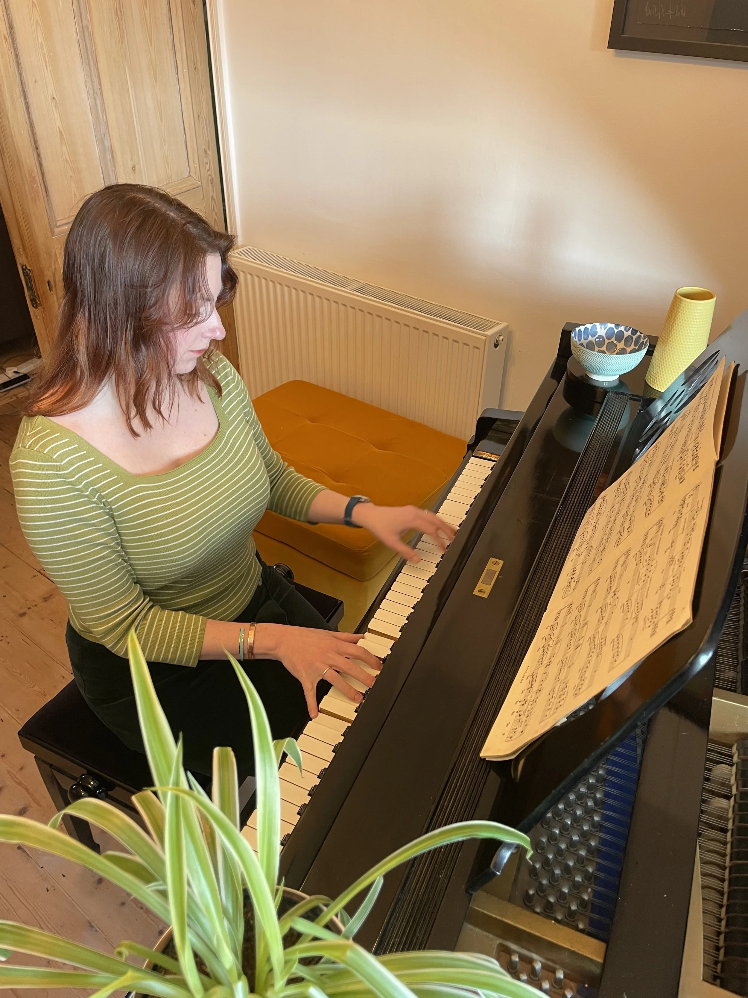 A woman with brown hair playing a black upright piano in a room, with sheet music on the music stand, a bowl, a yellow container, and some plants nearby.