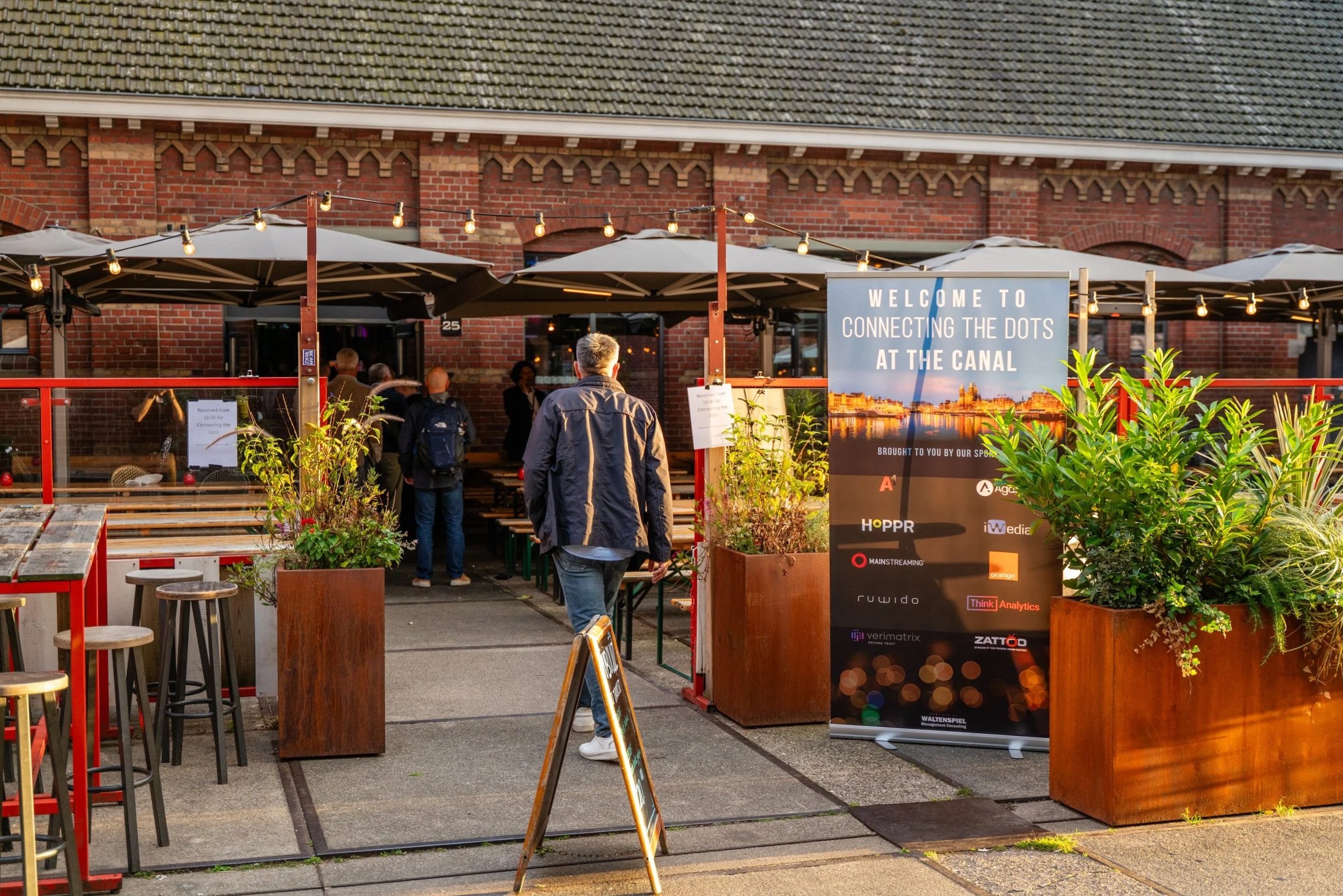 People entering a restaurant or cafe, with a sign saying "Welcome to Connecting the Dots at the Canal" and potted plants outside.