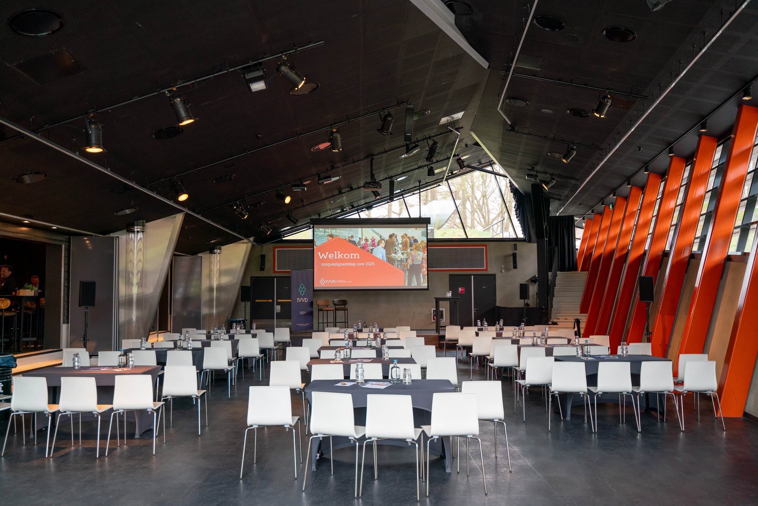 Conference room set up with tables and chairs facing a stage with a large screen displaying a welcoming message in Dutch, with social event photos, in a modern building with large windows and orange accents.