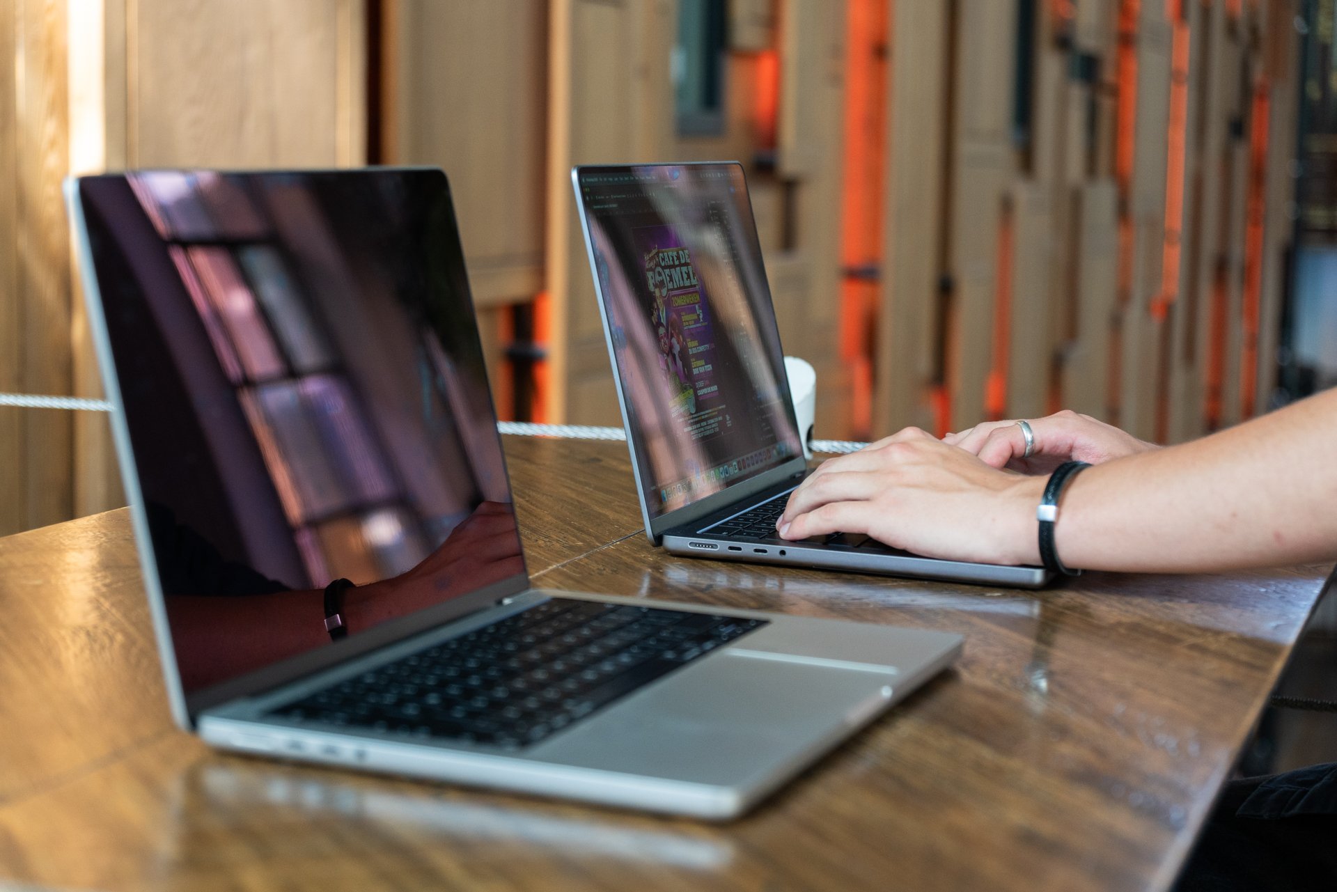 Two laptops on a wooden table, one open and one closed, with a person's hands typing on the open laptop in the background.