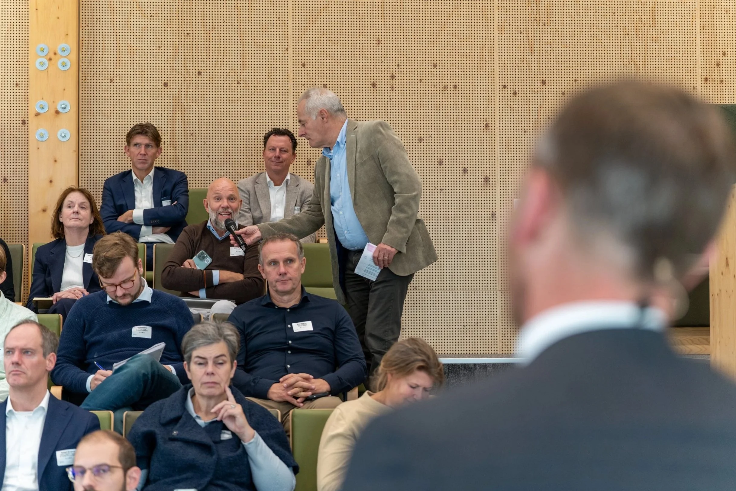 A man in a tan jacket holding a microphone asks a question at a conference with an audience seated in green chairs, some taking notes, and a speaker's podium visible in the foreground.