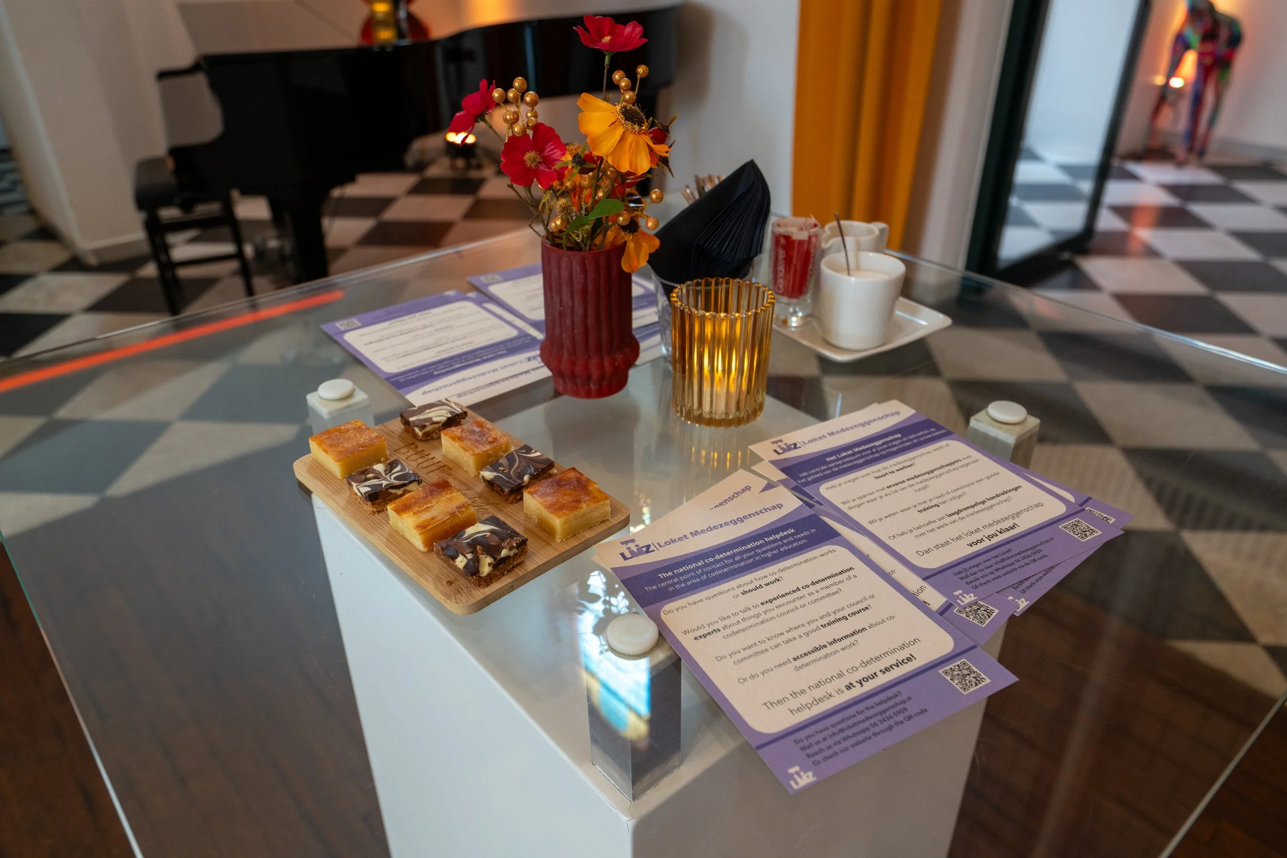 A glass table with a red vase holding red, orange, and pink flowers, a golden candle holder, two white cups with teaspoon and red drink, a tray with a napkin, brochures, and a wooden board with assorted desserts in chocolate and caramel flavors. The background shows a checkered floor, a black piano, and a yellow curtain.