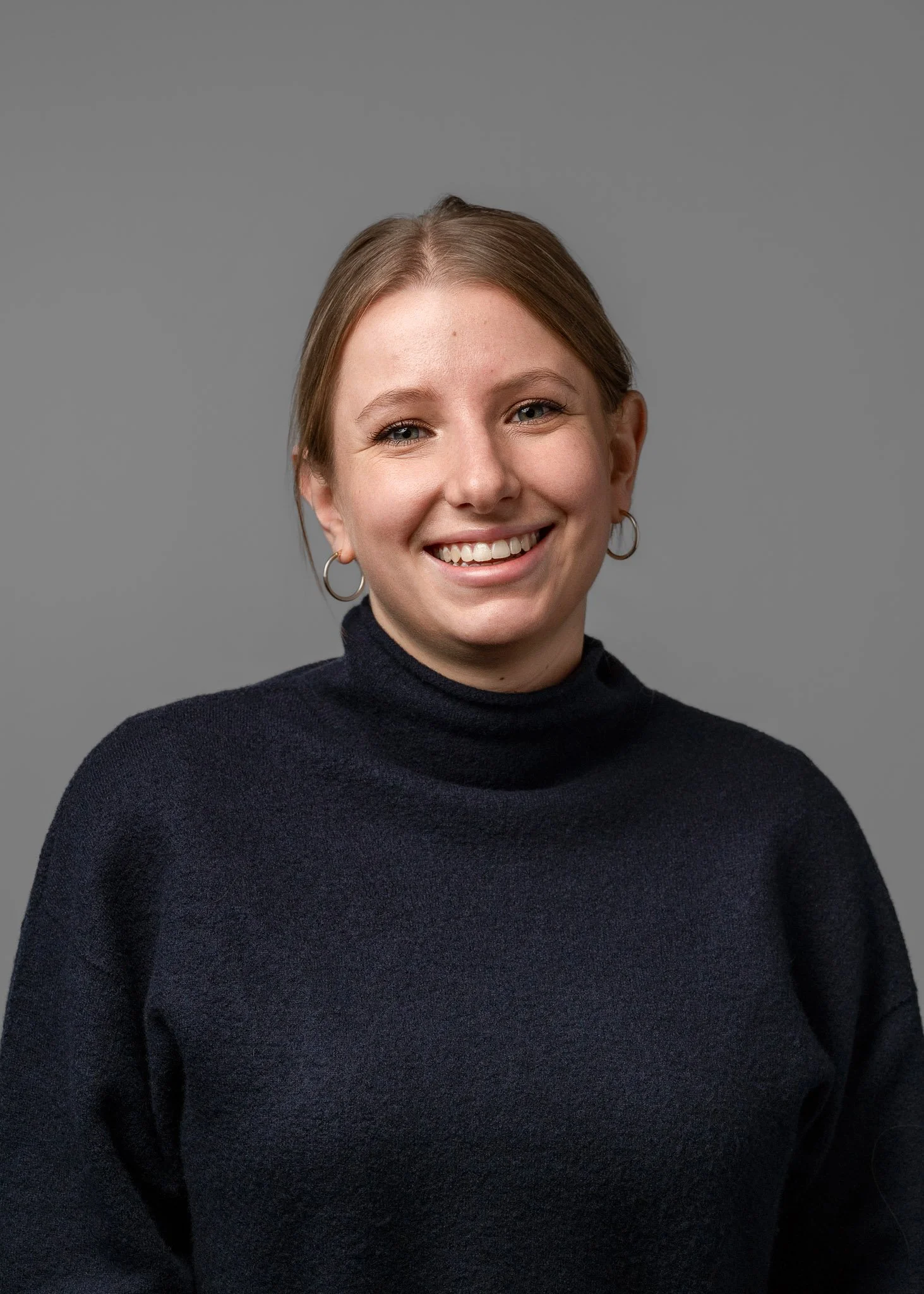 Portrait of a woman smiling with light makeup, wearing hoop earrings and a dark turtleneck sweater, against a gray background.
