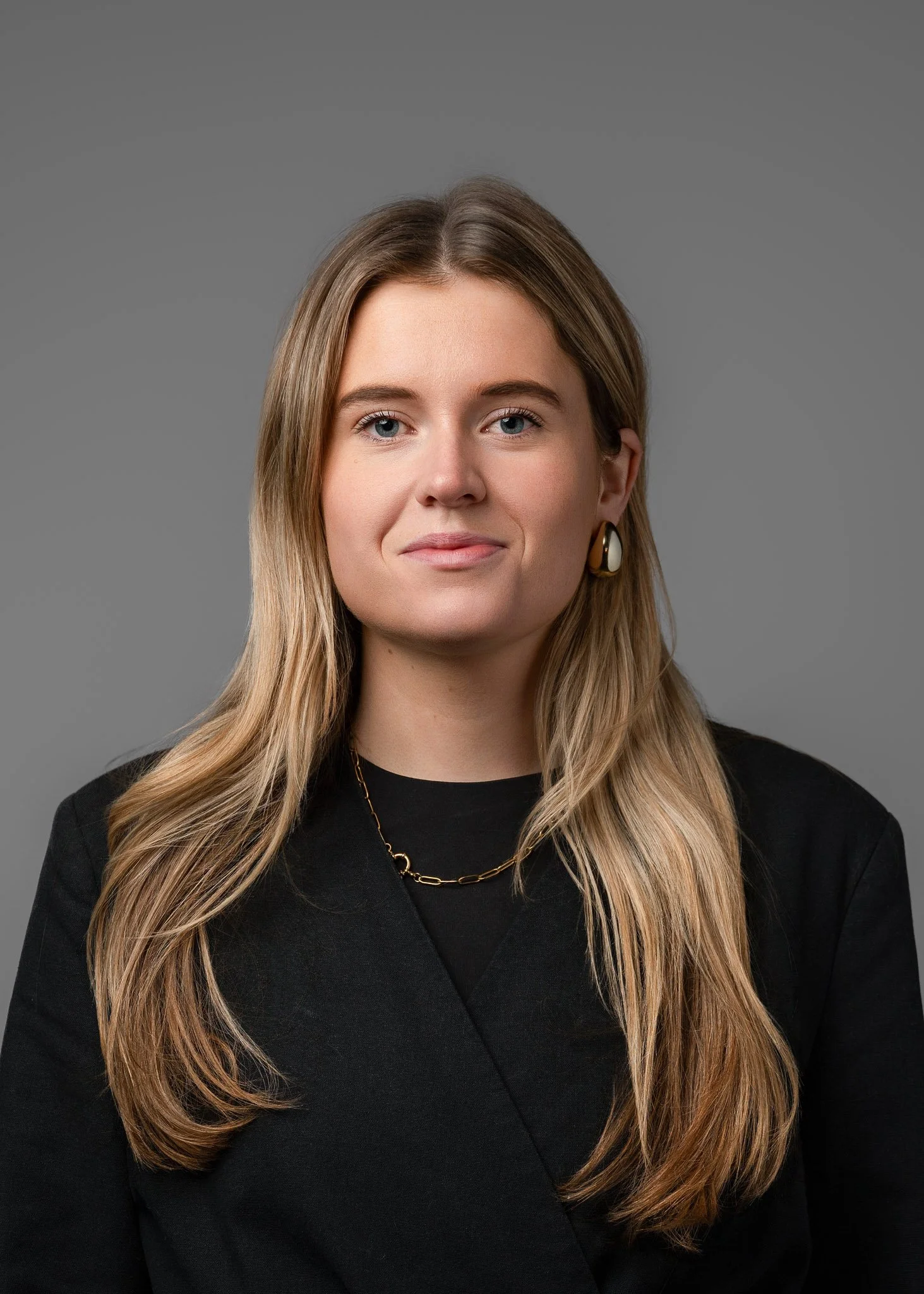A woman with long wavy blonde hair wearing a black blazer, gold chain necklace, and earrings standing against a gray background.