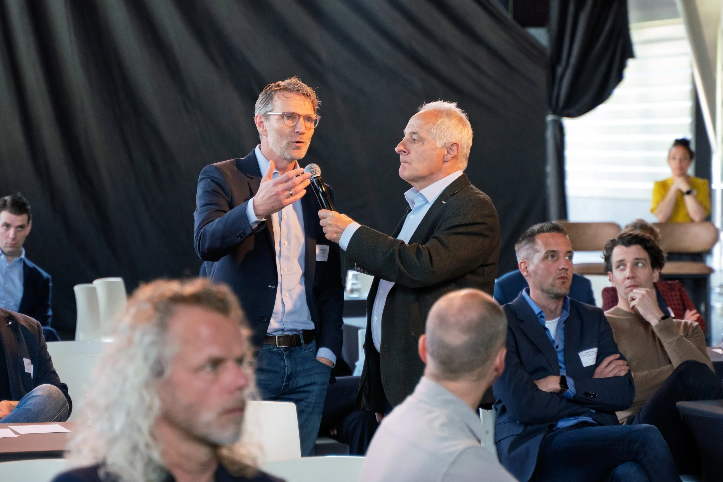 A man with a microphone is speaking while another man holds the microphone during a discussion at a conference, with an audience listening.