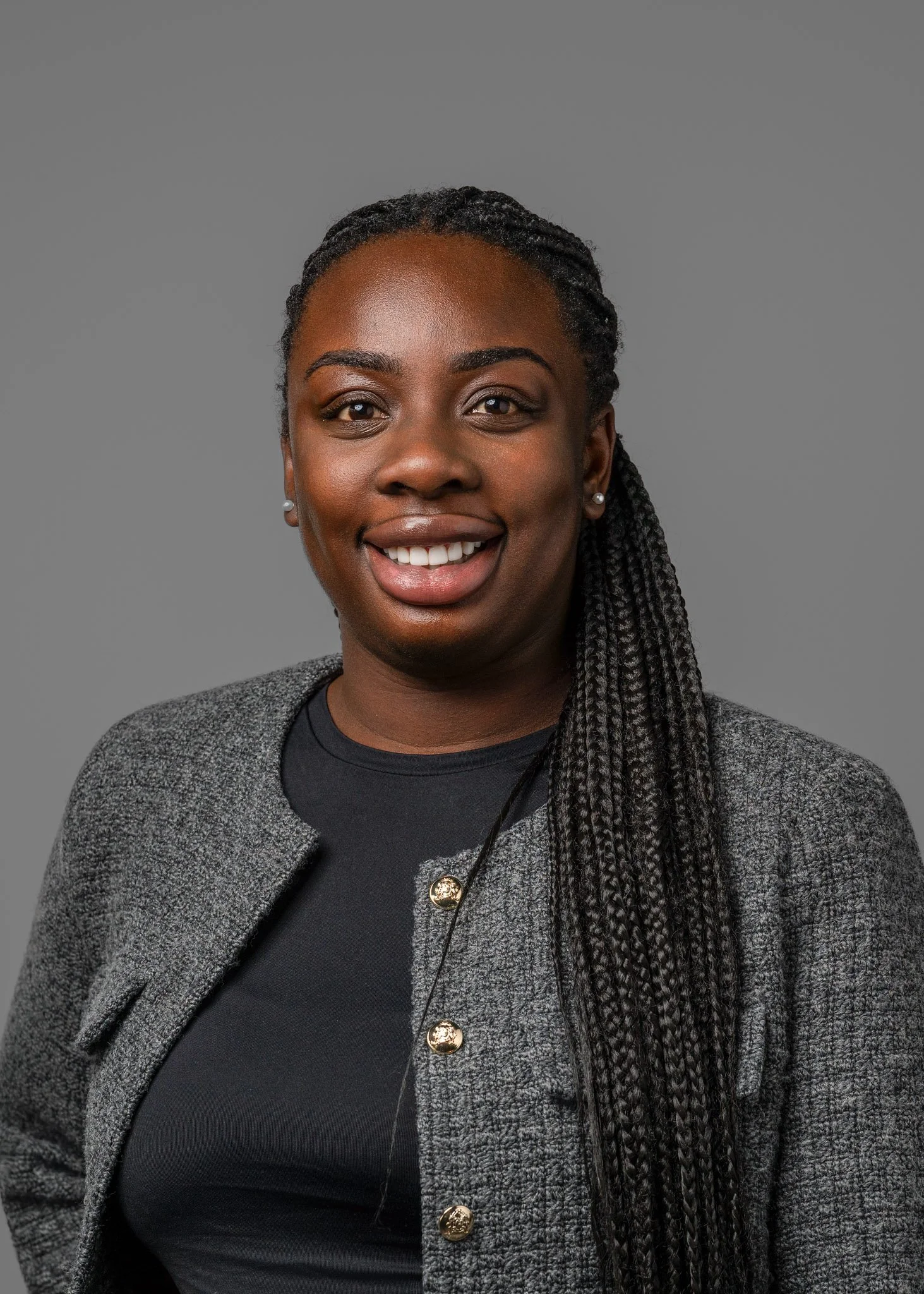 A smiling woman with long braided hair, wearing a black top and a gray blazer, posing against a gray background.