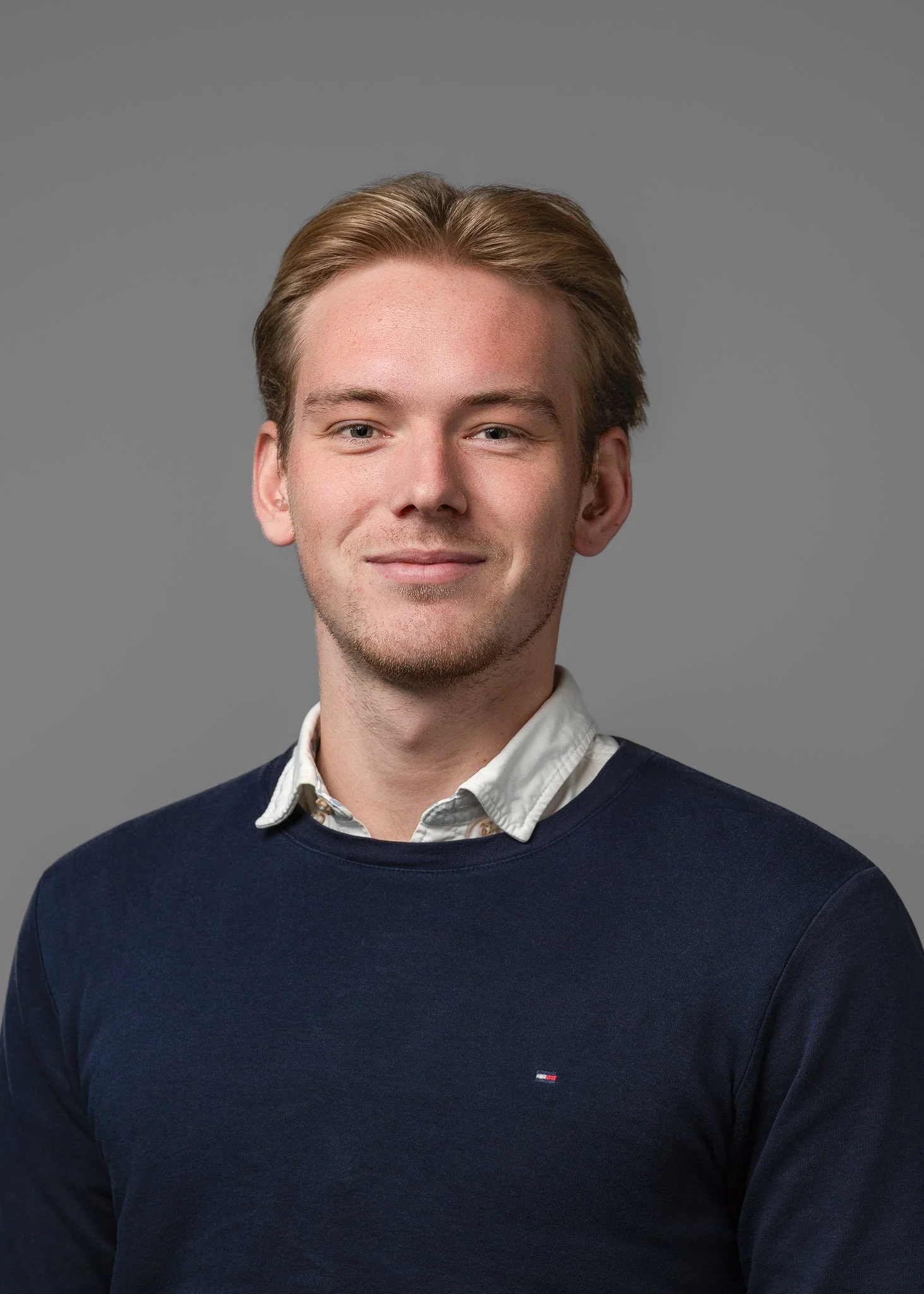 A young man with light skin, brown hair, and blue eyes. Wearing a white collared shirt underneath a navy sweater with a small logo on the chest, against a plain gray background.