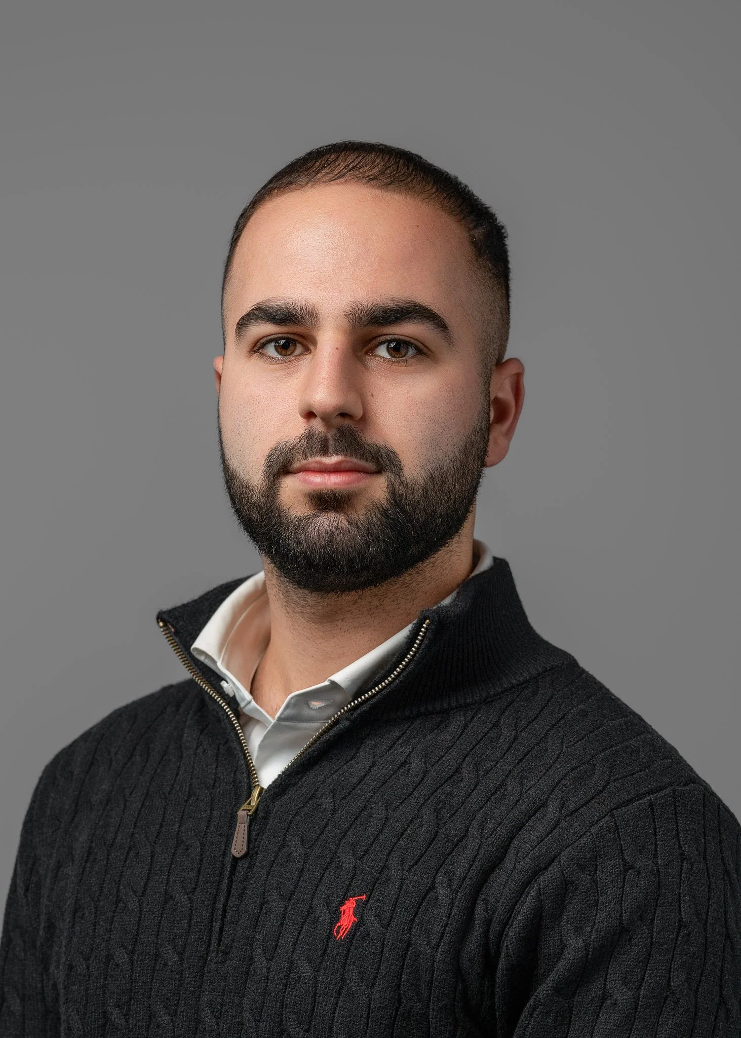 Headshot of a man with a beard and short hair, wearing a black zip-up sweater with a red Polo logo and a white collared shirt underneath, against a gray background.