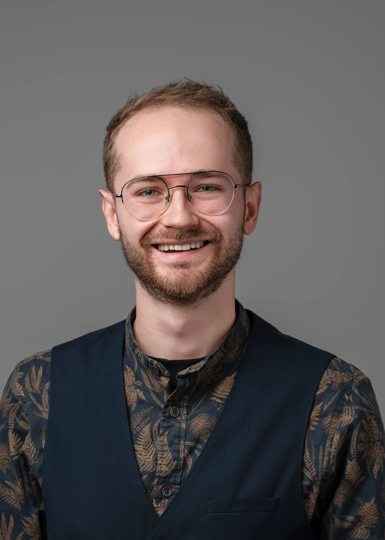 A smiling young man with glasses, a beard, and short light brown hair, wearing a black vest over a patterned shirt, standing against a plain gray background.