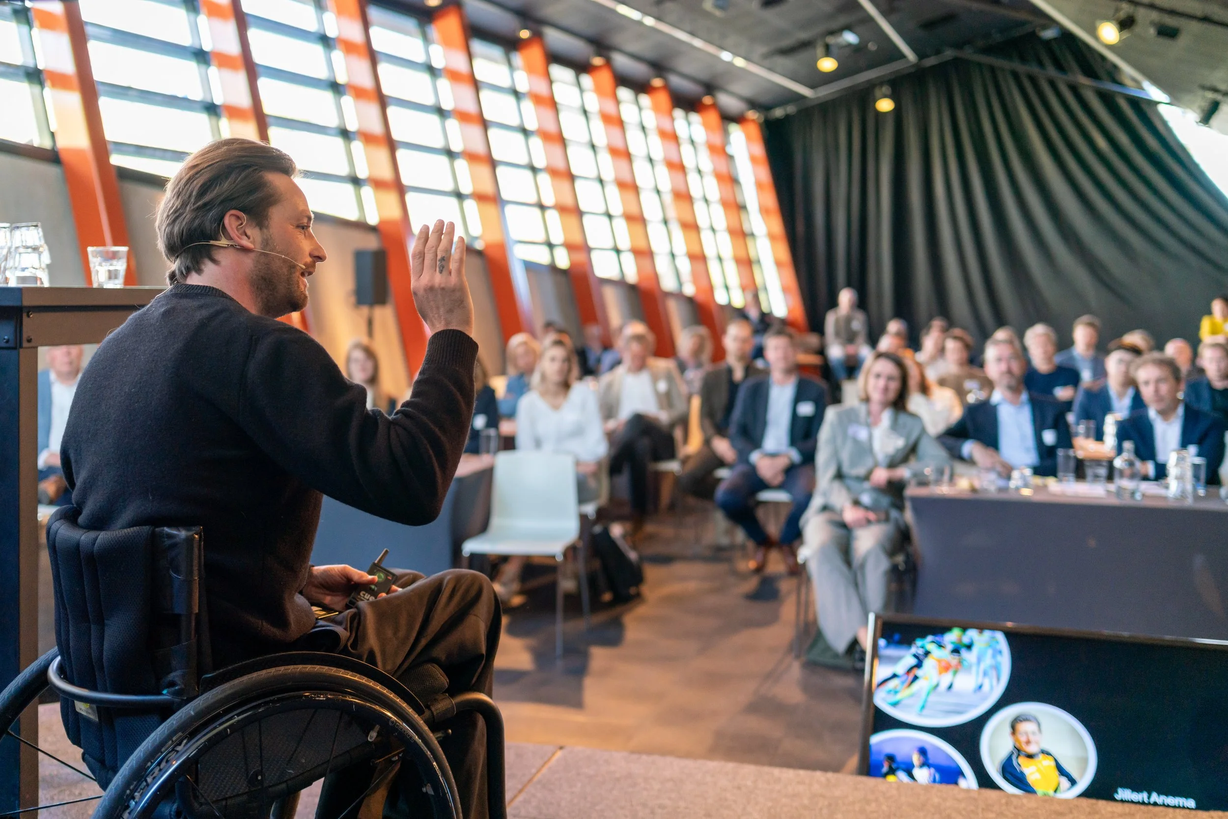 A man in a wheelchair giving a presentation to an audience in a conference room with large windows and black curtains.