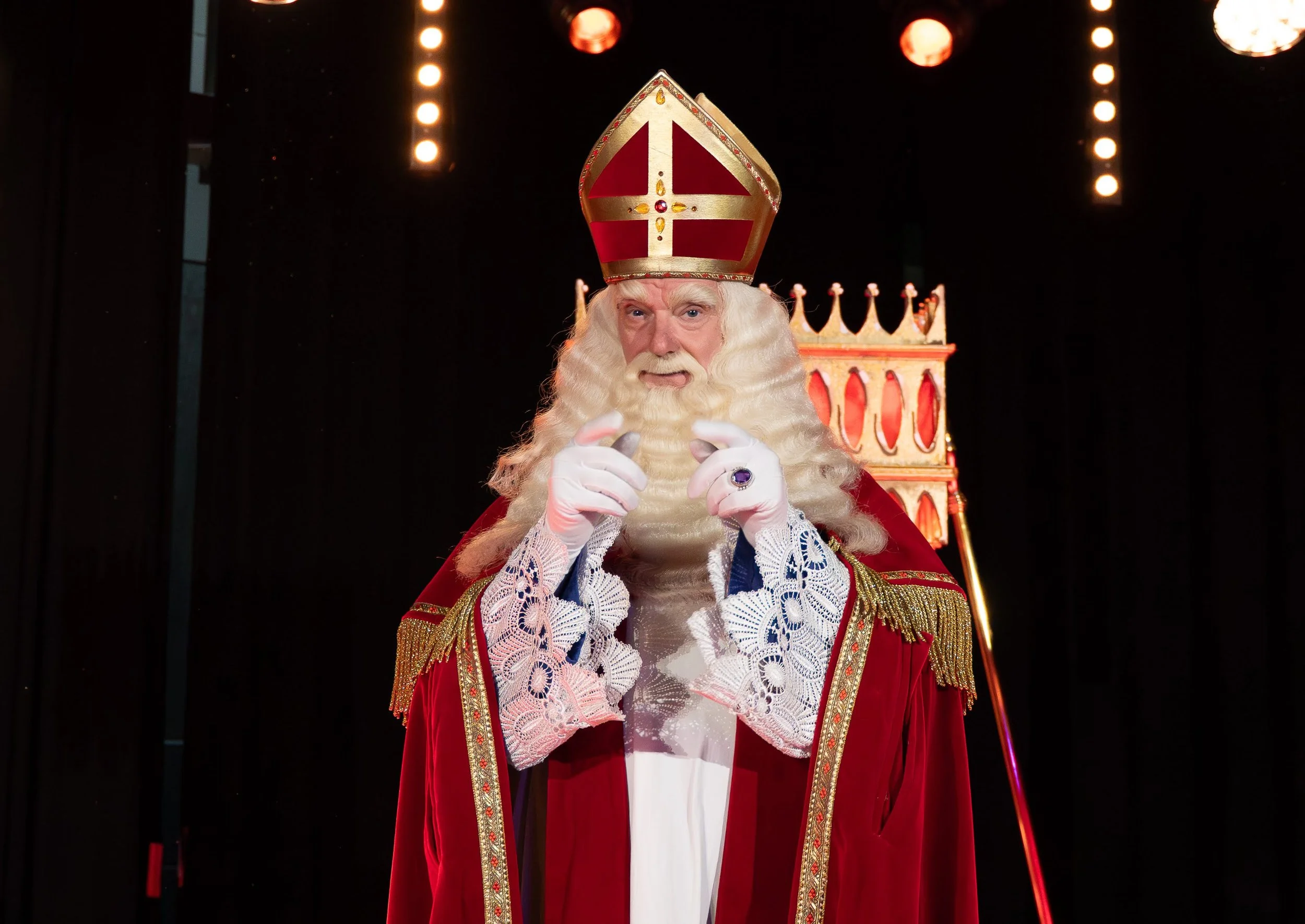 A man dressed as a bishop or saint sitting on a throne with a tall, ornate crown, wearing a red robe with gold and white lace details, pointing with both hands. The background is dark with bright lights overhead.