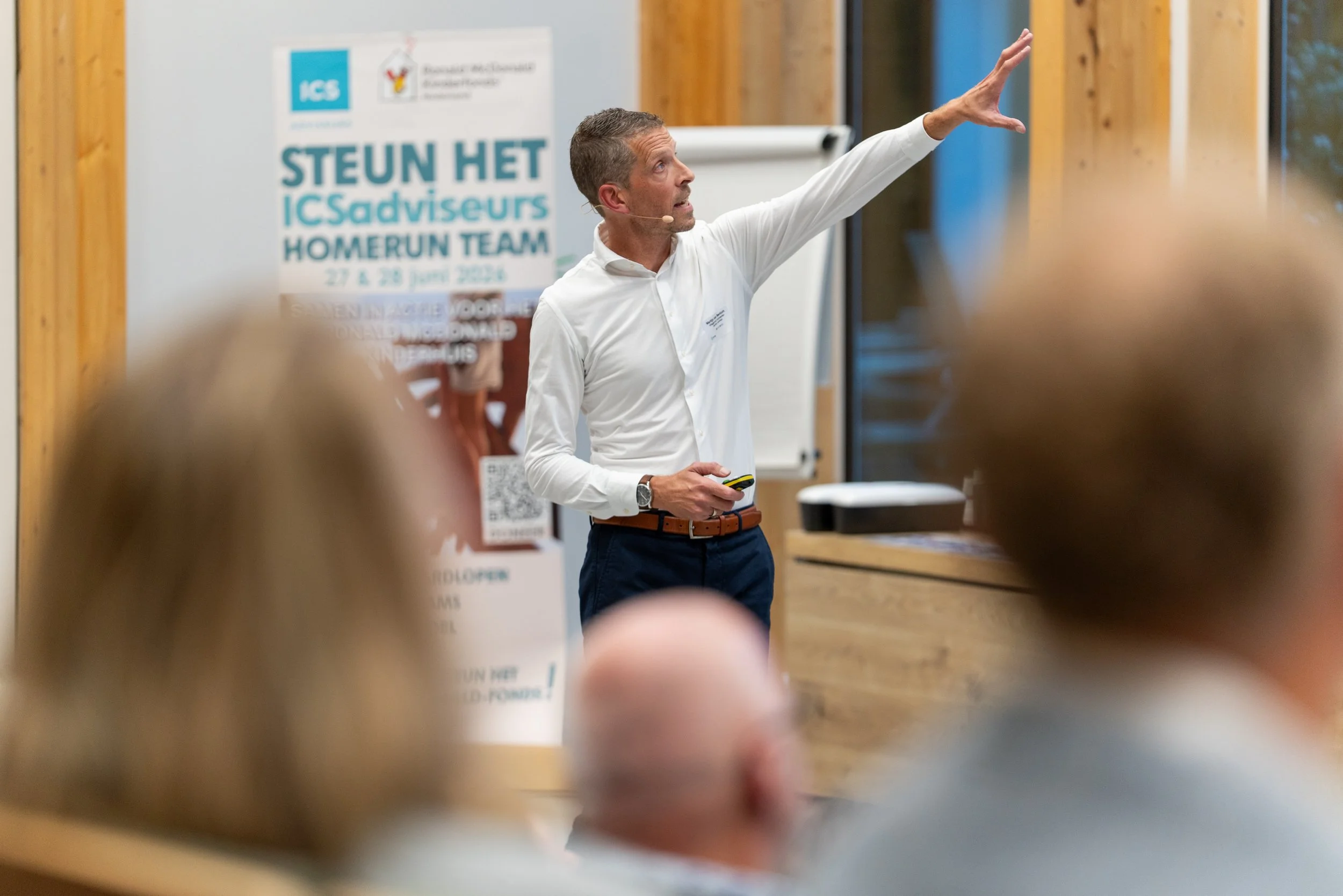 A man in a white shirt giving a presentation to an audience with people in the foreground. There is a poster behind him with text in Dutch and English.