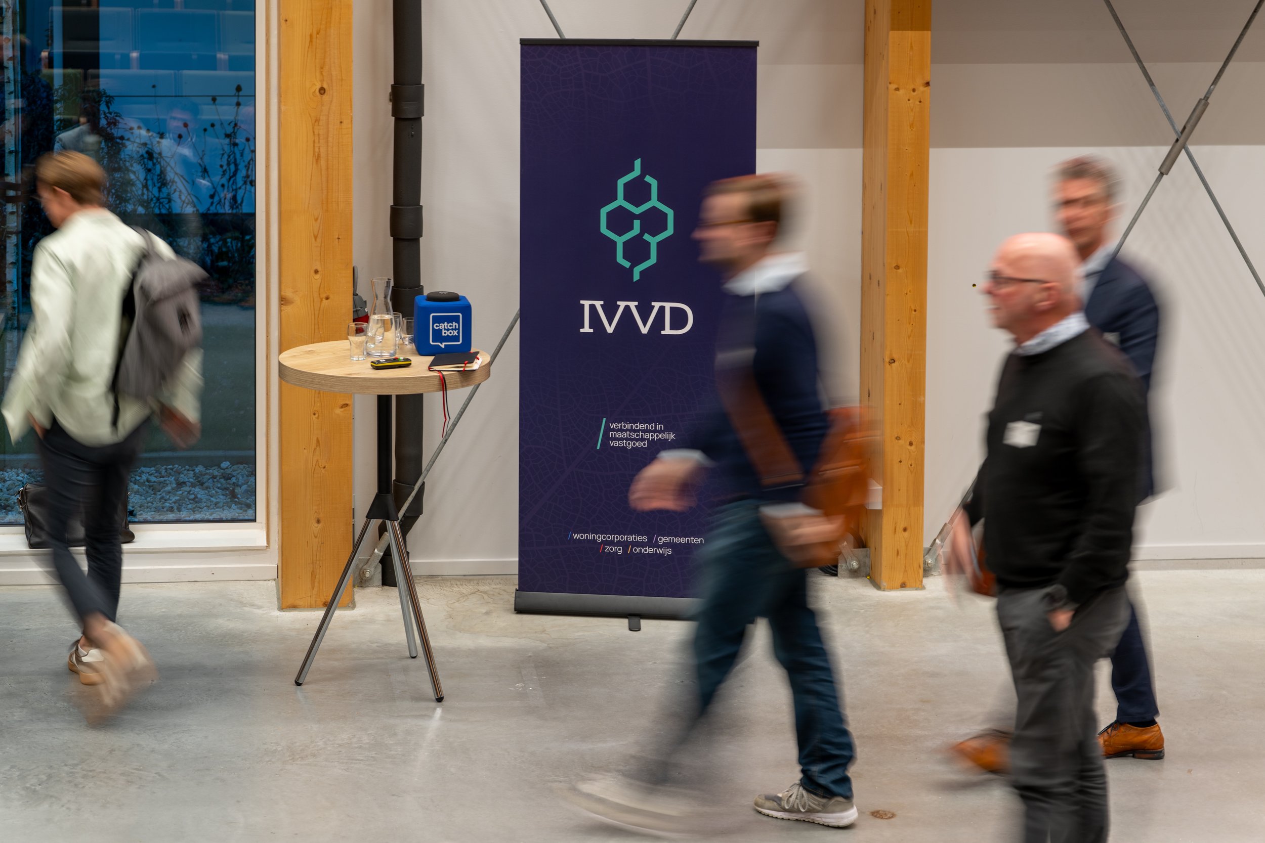 People walking past a large purple IVVD banner at an indoor event, with a small round table holding a blue speaker, two glasses, and a red and yellow item nearby.