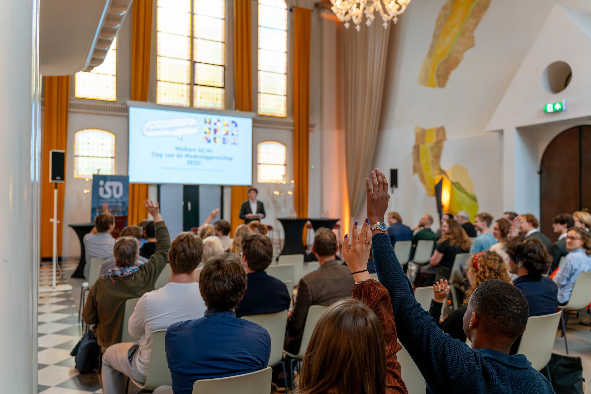 Audience participating in a conference with several people raising their hands, a presenter standing at the front, a large screen displaying a welcome message, and a historic, ornately decorated room with high stained-glass windows and yellow curtains.