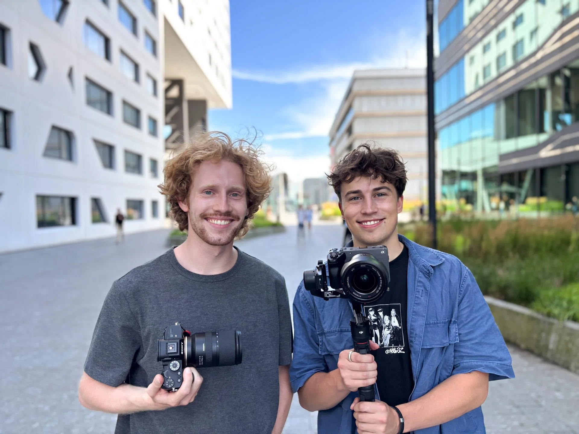 Two young men smiling outdoors, one holding a camera in one hand and the other holding a stabilizer with a camera, in an urban area with modern buildings.