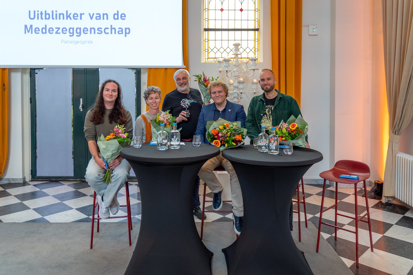 Group of five people at a panel discussion event, holding flowers and awards, seated behind black round tables with water bottles and glasses in a room with a large screen, stained glass window, and curtains.