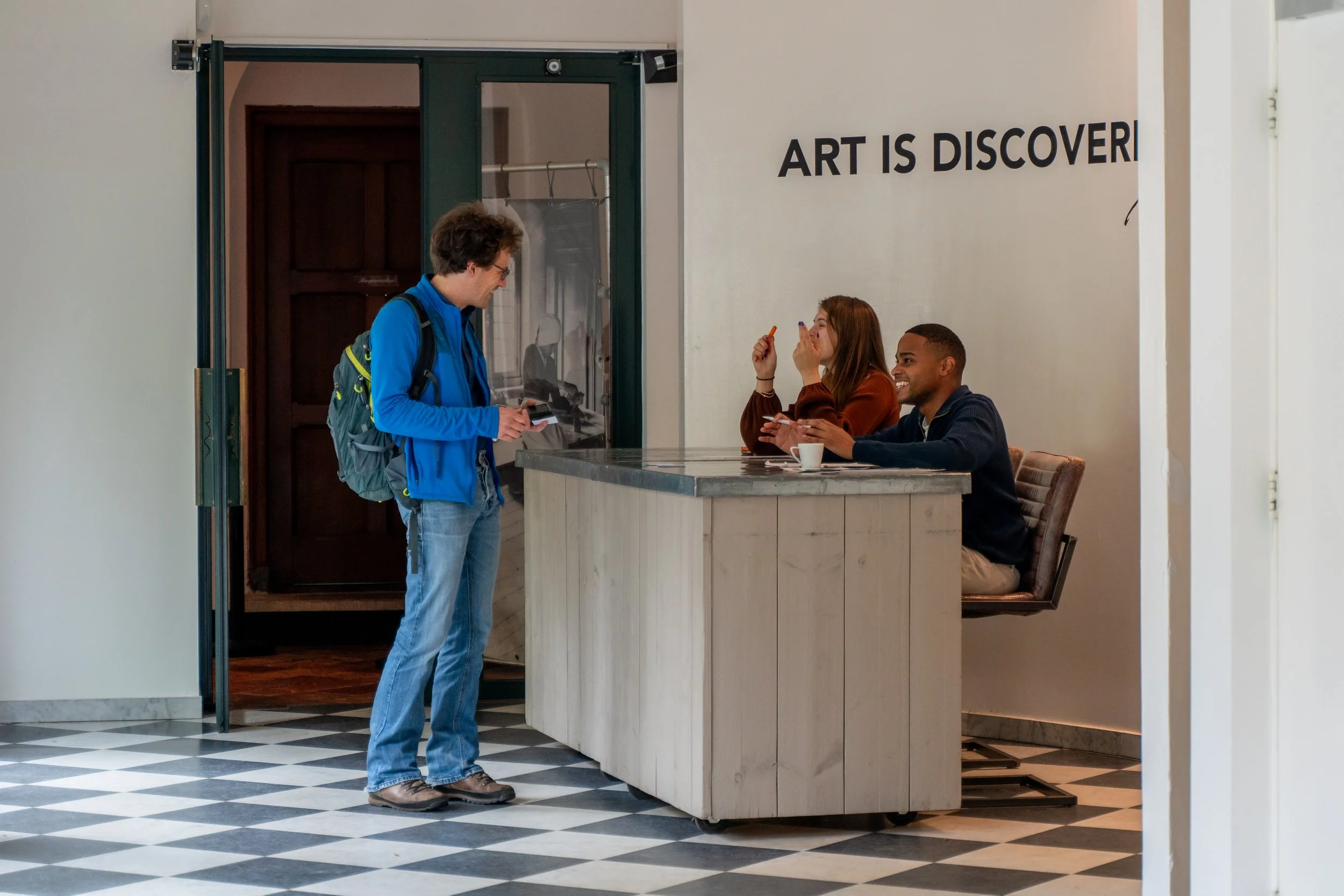 A young man with a backpack standing at a reception desk, talking to two women who are seated behind the desk. The women are smiling, and one is holding up a piece of paper. The background has a sign saying 'Art is Discovery'.