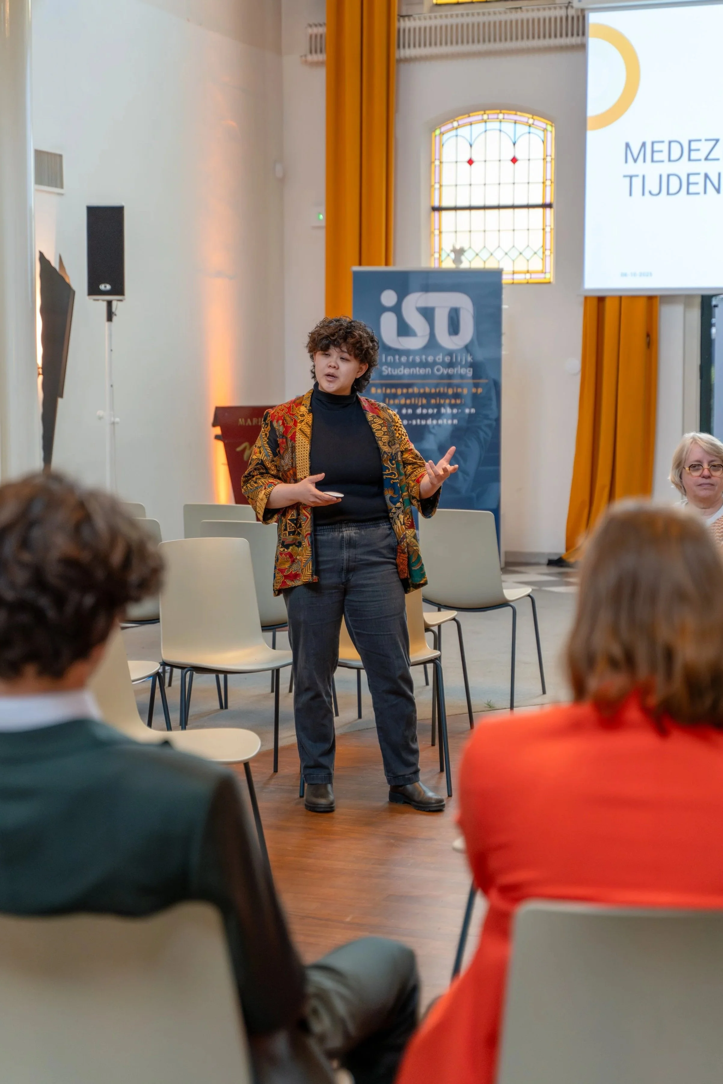 A woman giving a presentation in front of a small audience in a room with large windows and orange curtains. A large screen displays a slide with text in Dutch, and a vertical banner with Danish text is visible in the background.