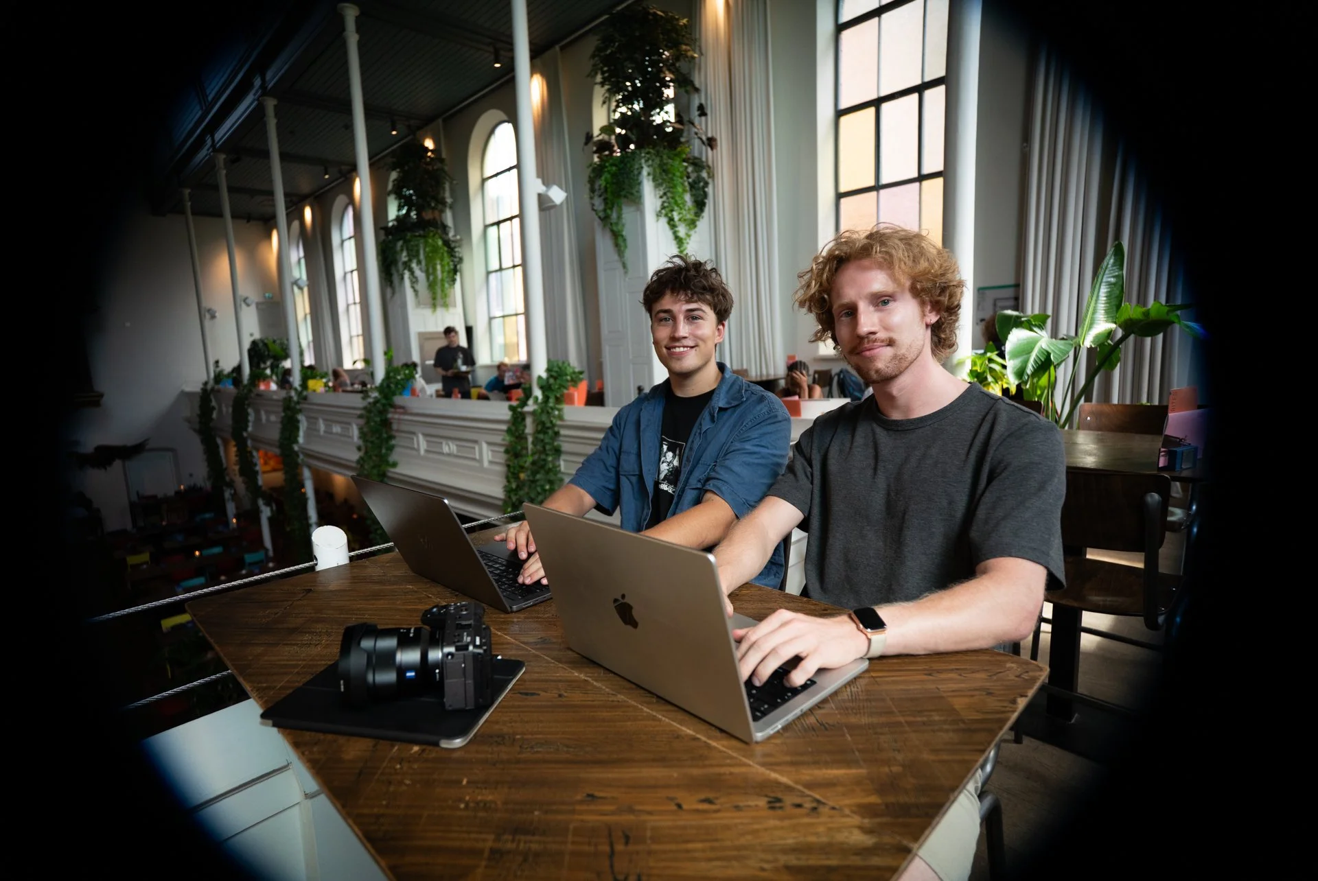 Two young men sitting at a wooden table in a bright, spacious cafe or co-working space, working on laptops, with a camera and some other items on the table, large windows, and green plants in the background.