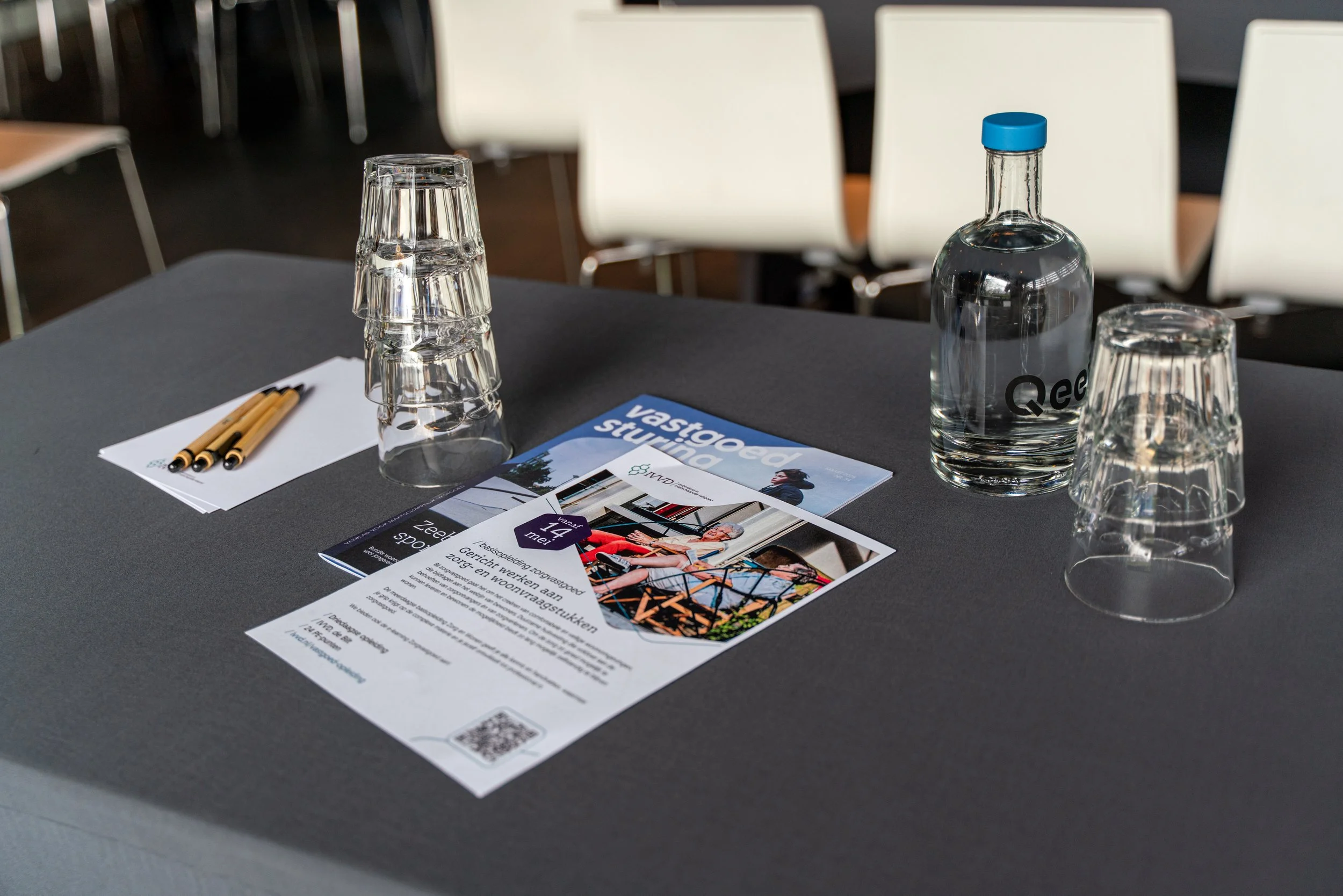 Table setup with three stacked glasses, a bottle of water, pens, and informational brochures, with chairs in the background.