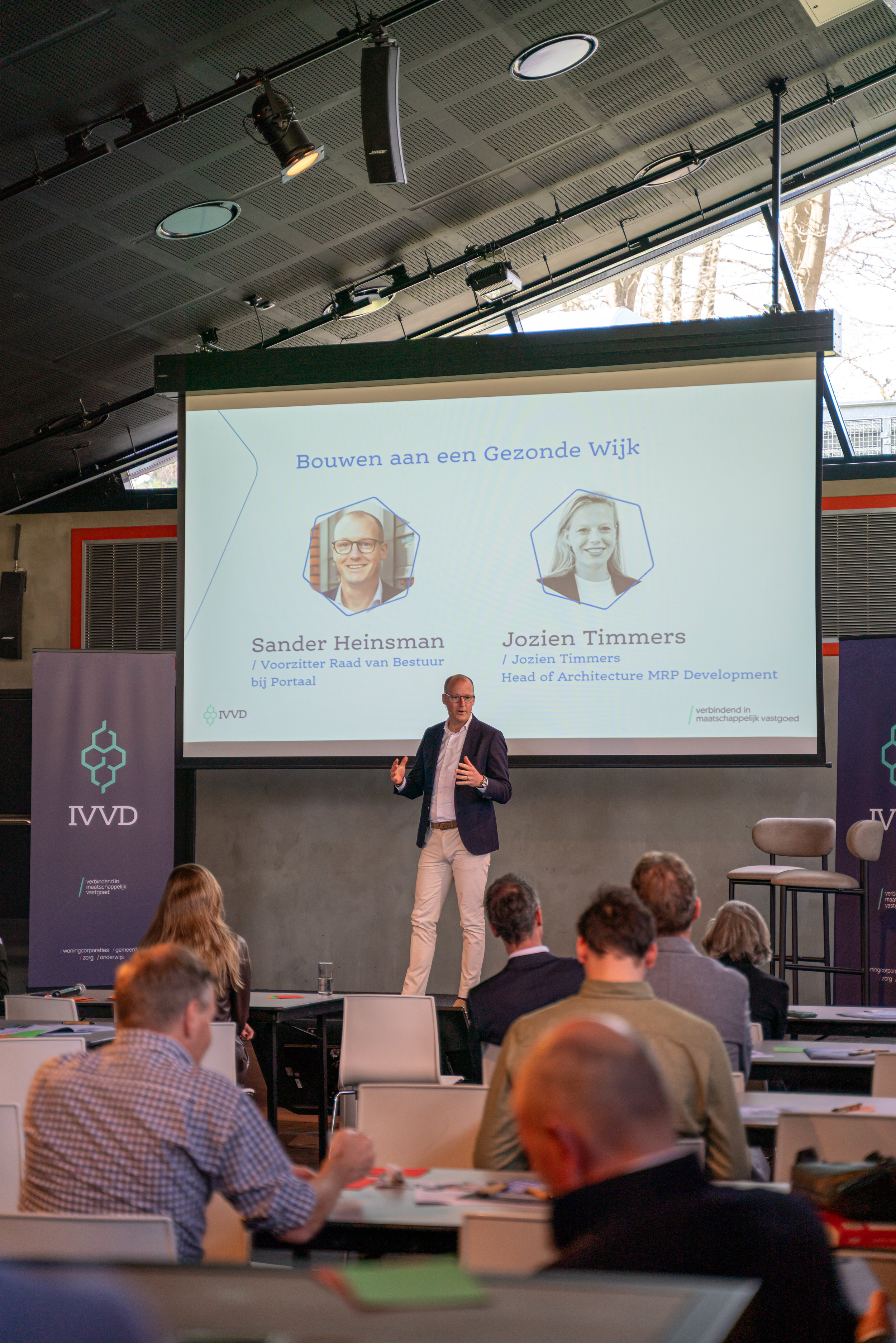 A man giving a presentation in a conference room with a large screen behind him displaying a slide titled 'Bouwen aan een Gezonde Wijk.' The slide features photos and names of Sander Heinsman and Jozien Timmers. There are attendees seated at tables listening to the presentation, with purple IVVD banners on either side of the stage.