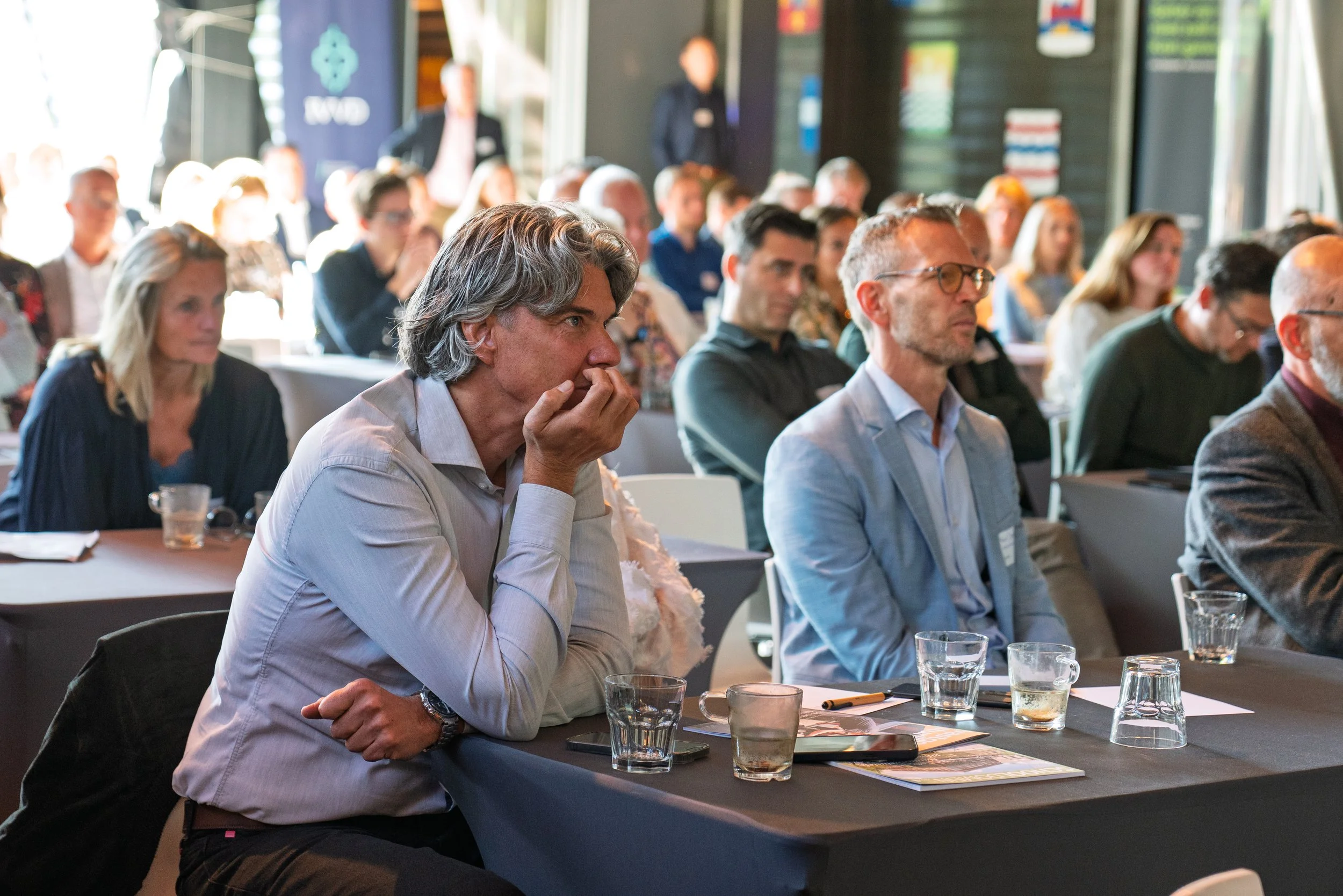Audience listening attentively at a conference or seminar in a dimly lit room with dark walls and flags in the background.