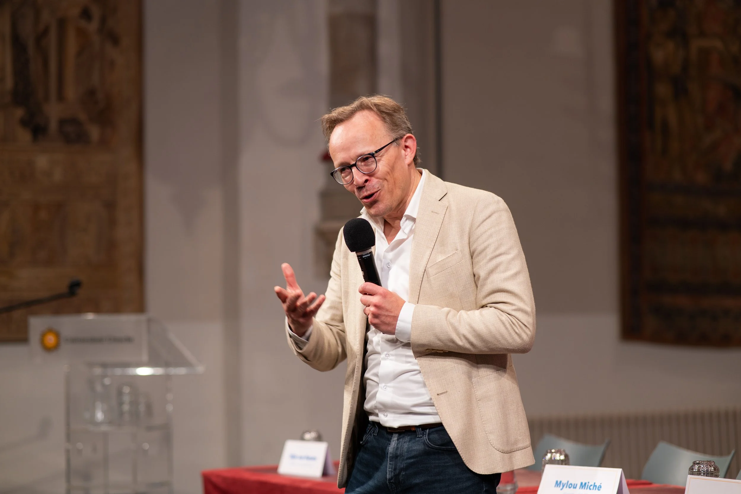 A man with glasses and light-colored blazer speaking into a microphone at a formal event.