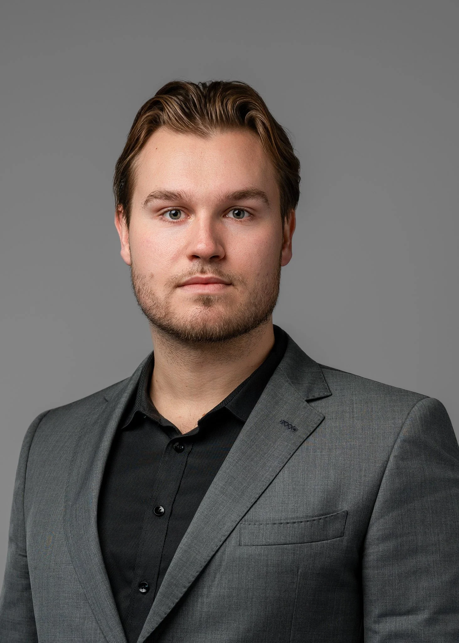 A young man with light skin and brown hair styled back, wearing a gray suit jacket over a black shirt, posing against a plain gray background.