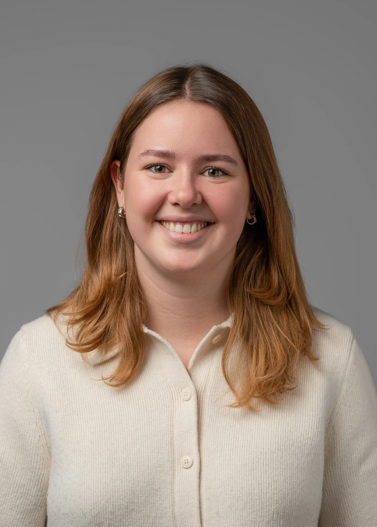 Portrait of a young woman with shoulder-length light brown hair, smiling, wearing a cream-colored button-up cardigan, earrings, against a gray background.