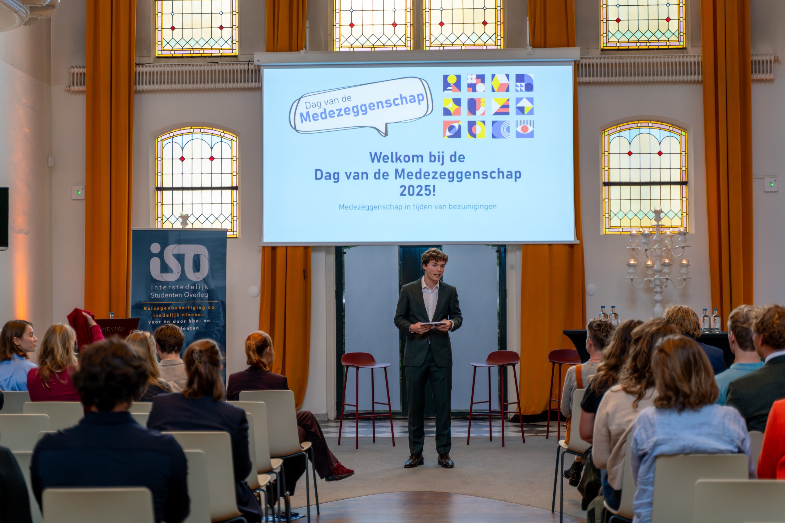 A young man in a suit giving a presentation during an event titled 'Dag van de Medezeggenschap 2025' in a decorated hall with stained glass windows, a large screen, and an audience seated facing the speaker.