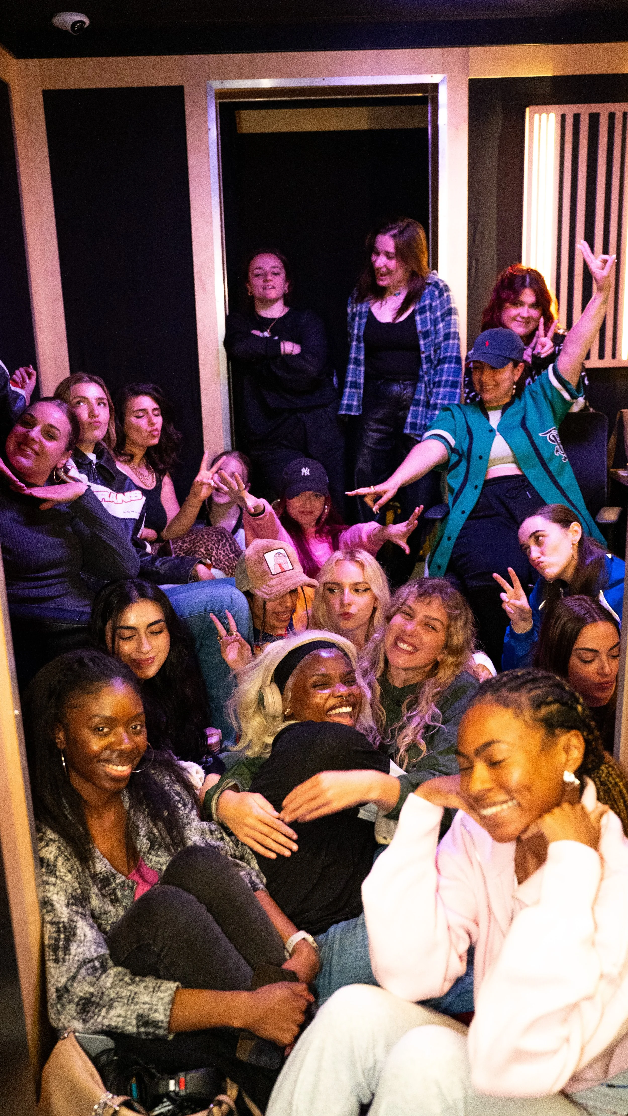Group of women and girls in a recording studio, some sitting and others standing, smiling and making playful gestures.
