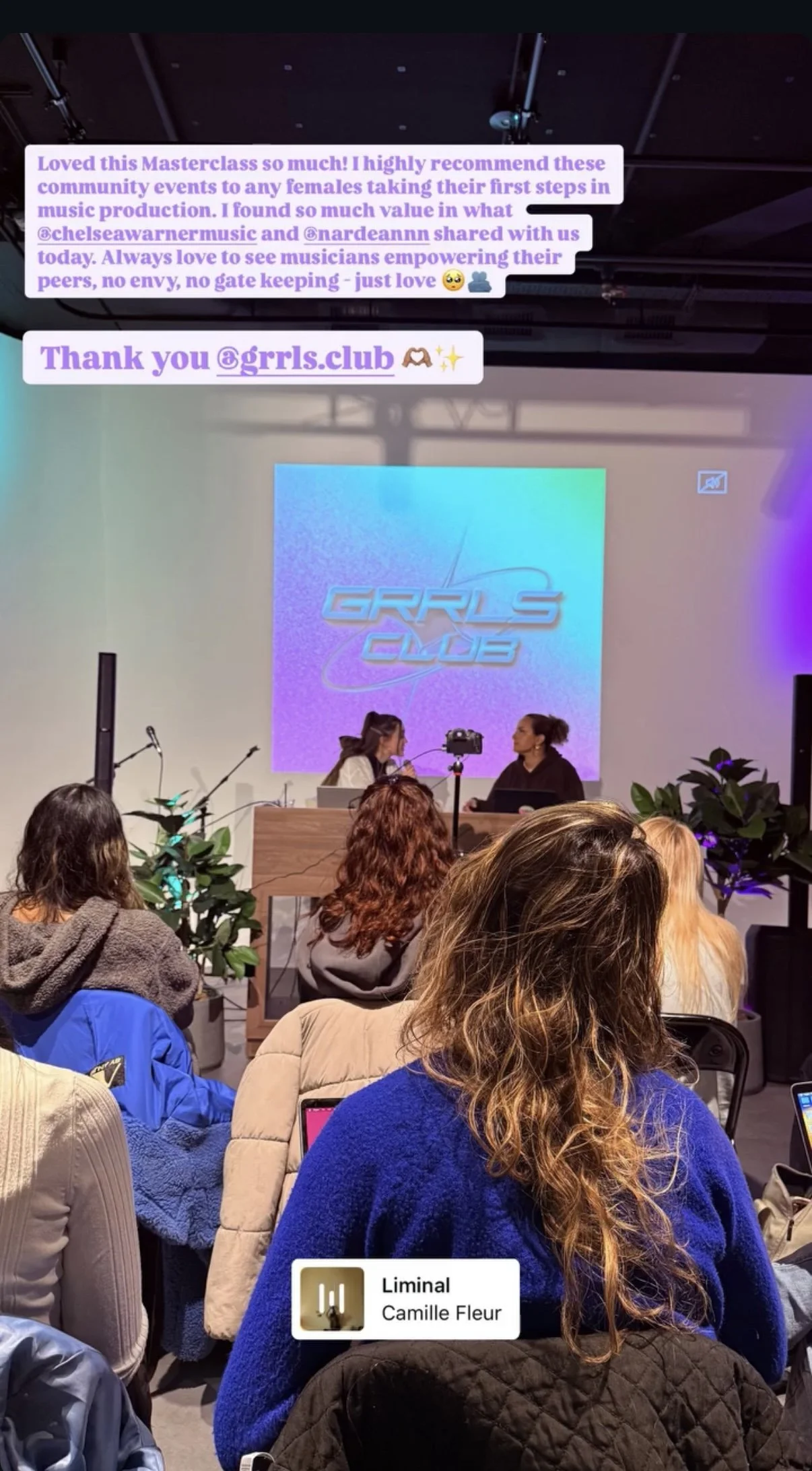 A group of women attending a music production masterclass called Grrls Club. Two women are on stage, engaged in conversation, with a large screen behind them displaying the Grrls Club logo. Audience members are seated facing the stage, some with laptops or phones, and plants are placed on the sides of the stage.