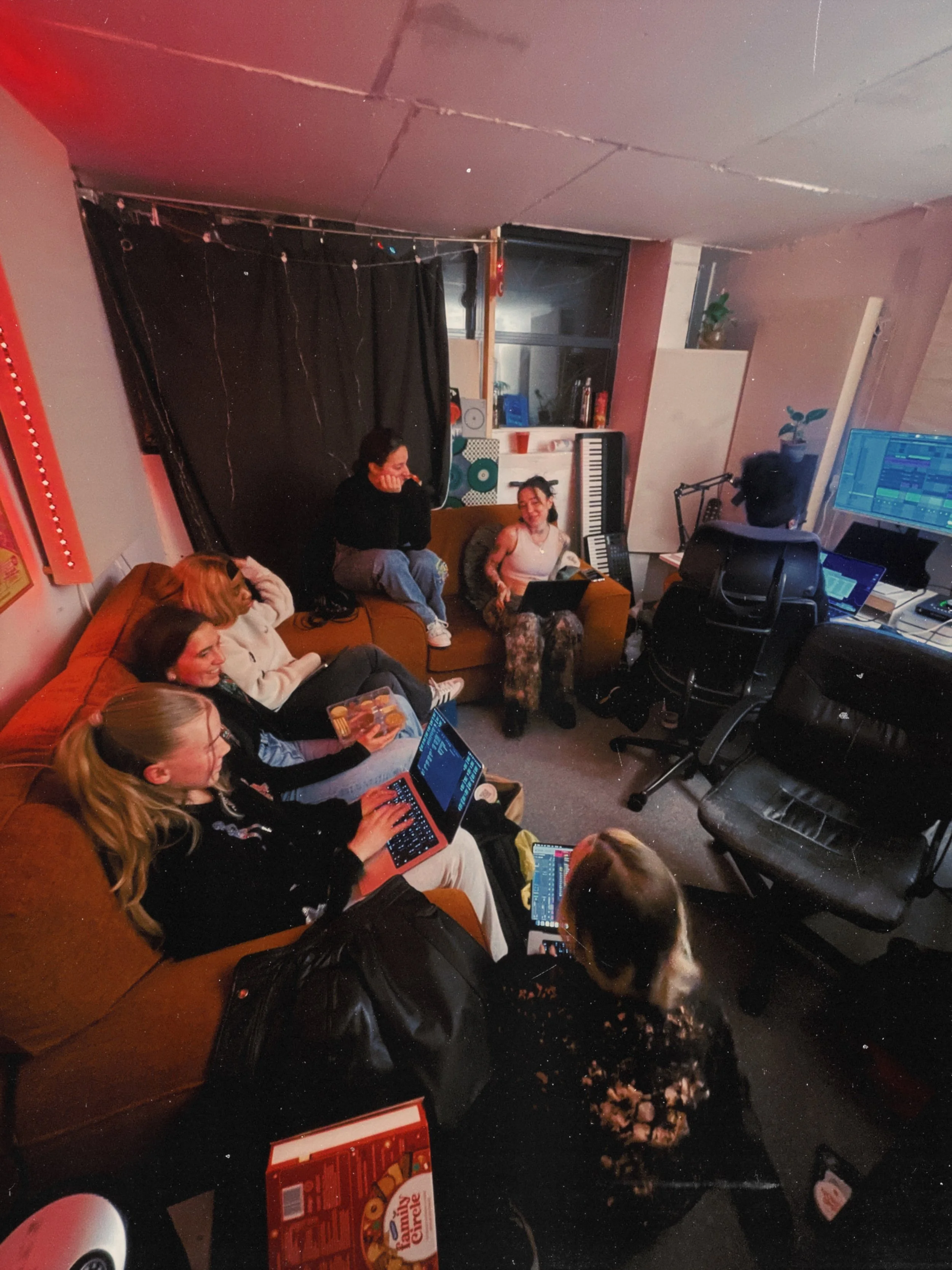 A group of friends gathered in a music studio or rehearsal space, sitting on couches and chairs, with laptops, a tablet, and a drum kit. There is a keyboard, a computer monitor, and various music equipment around them. The room has a black curtain, a window, and some decorative plants.