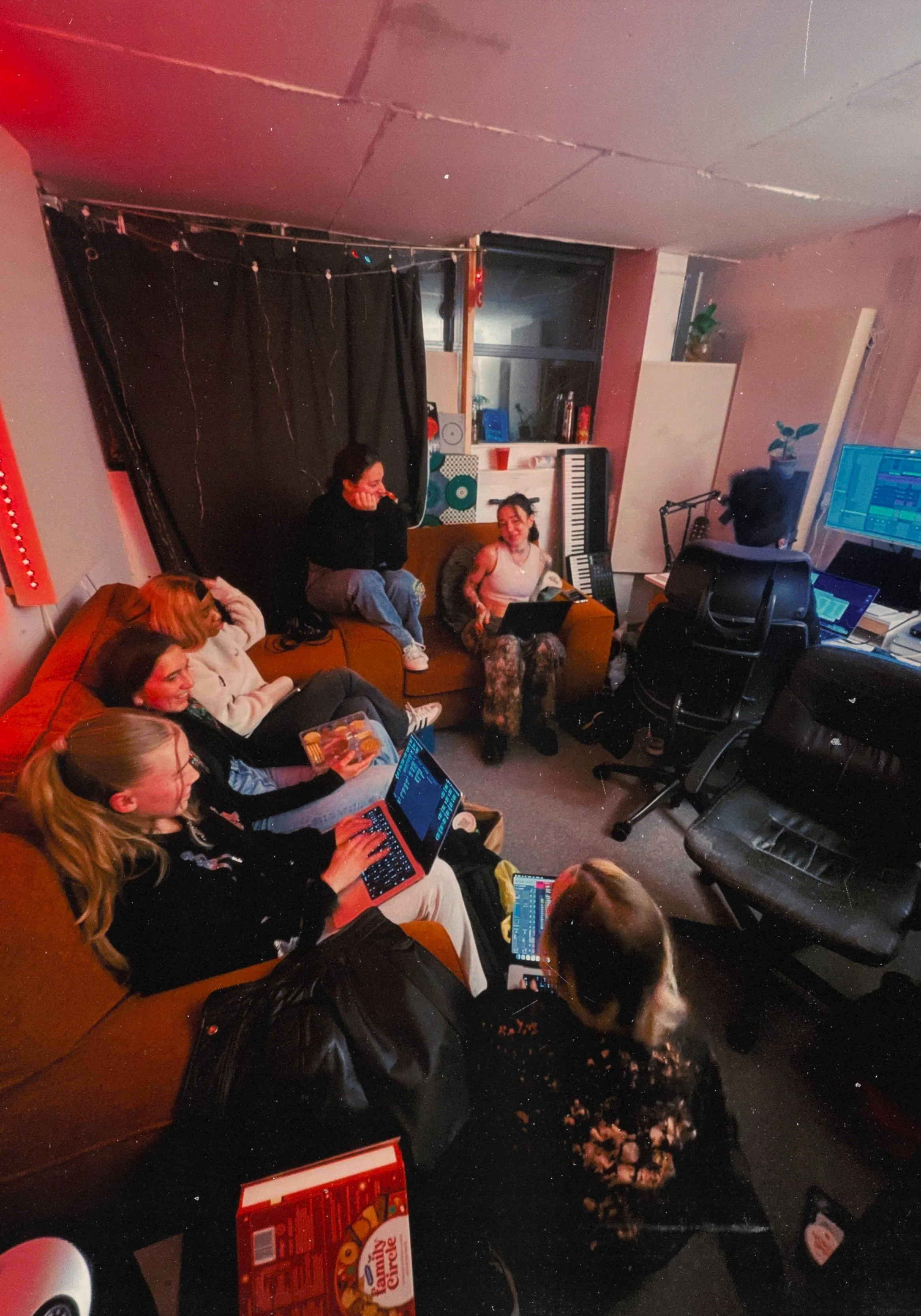 A group of young women in a cozy room with musical instruments and studio equipment, sitting on couches and chairs, working on laptops, and engaging in a music session or gathering.