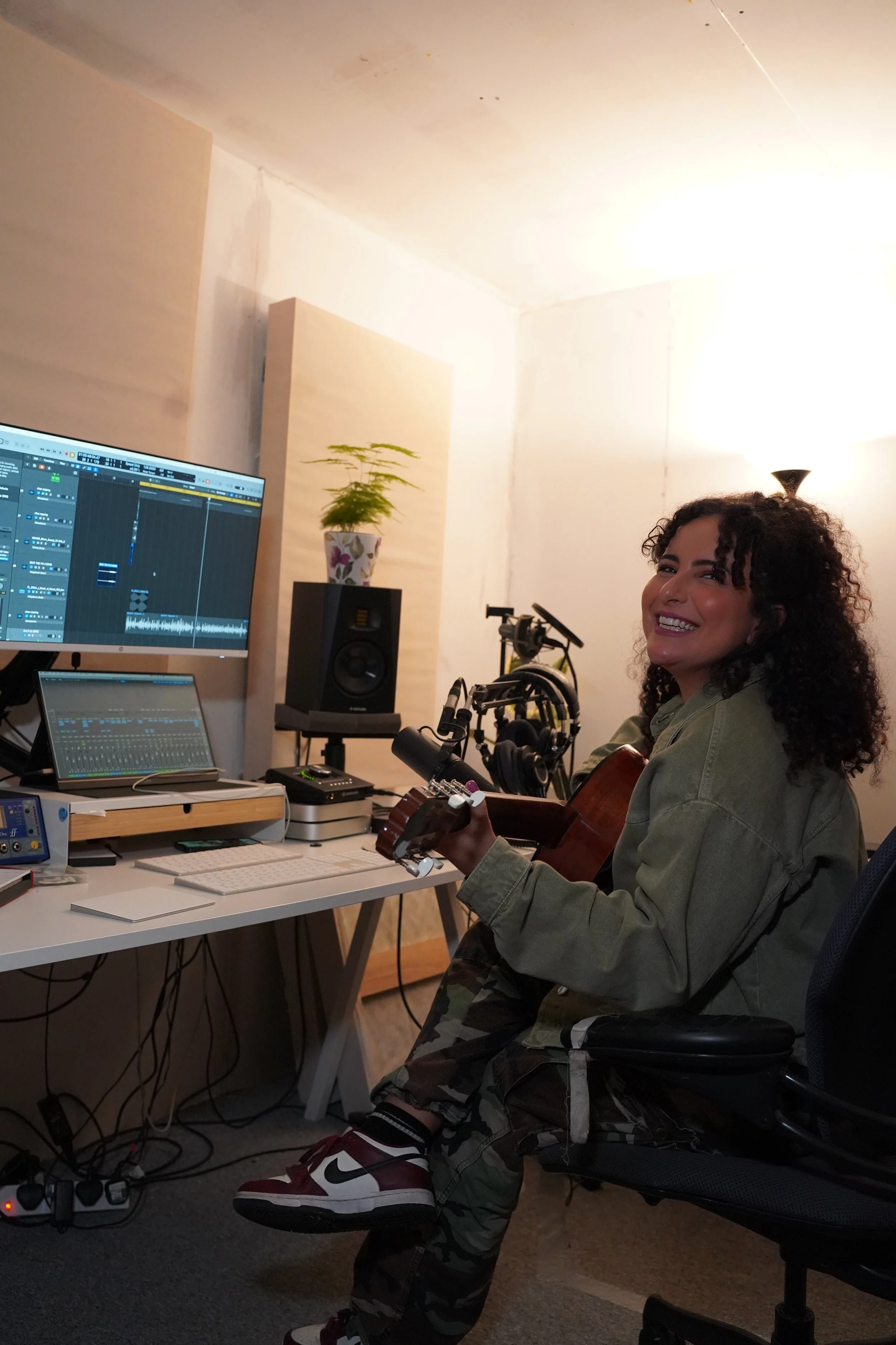A woman sitting in a home recording studio, holding a guitar and smiling at the camera. The studio has a computer with audio editing software, studio speakers, and various music equipment.