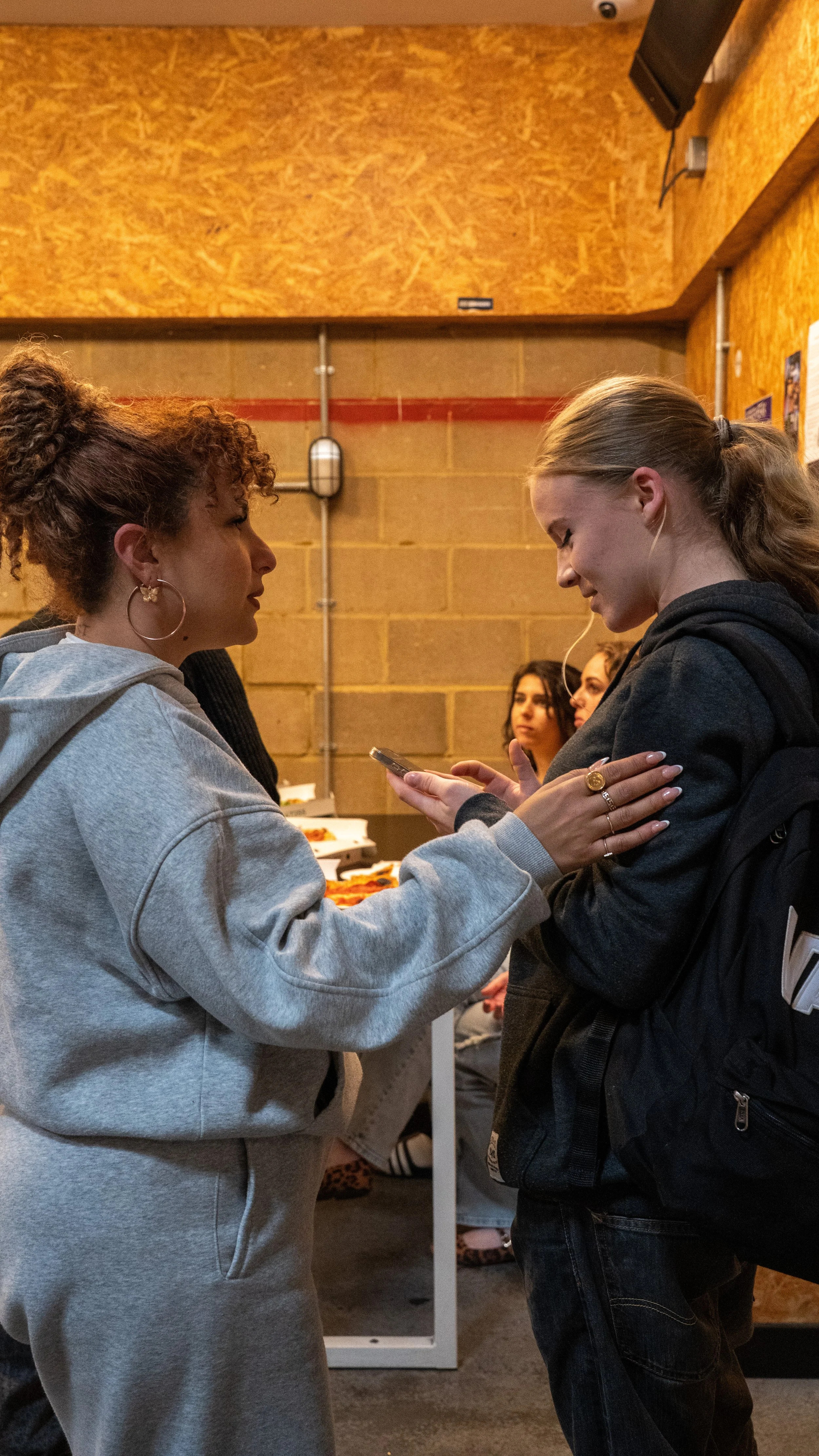 Two young women are standing face-to-face, engaging in a conversation, with one showing something on their phone to the other. In the background, three other women are sitting at a table with food, in a room with orange textured walls.